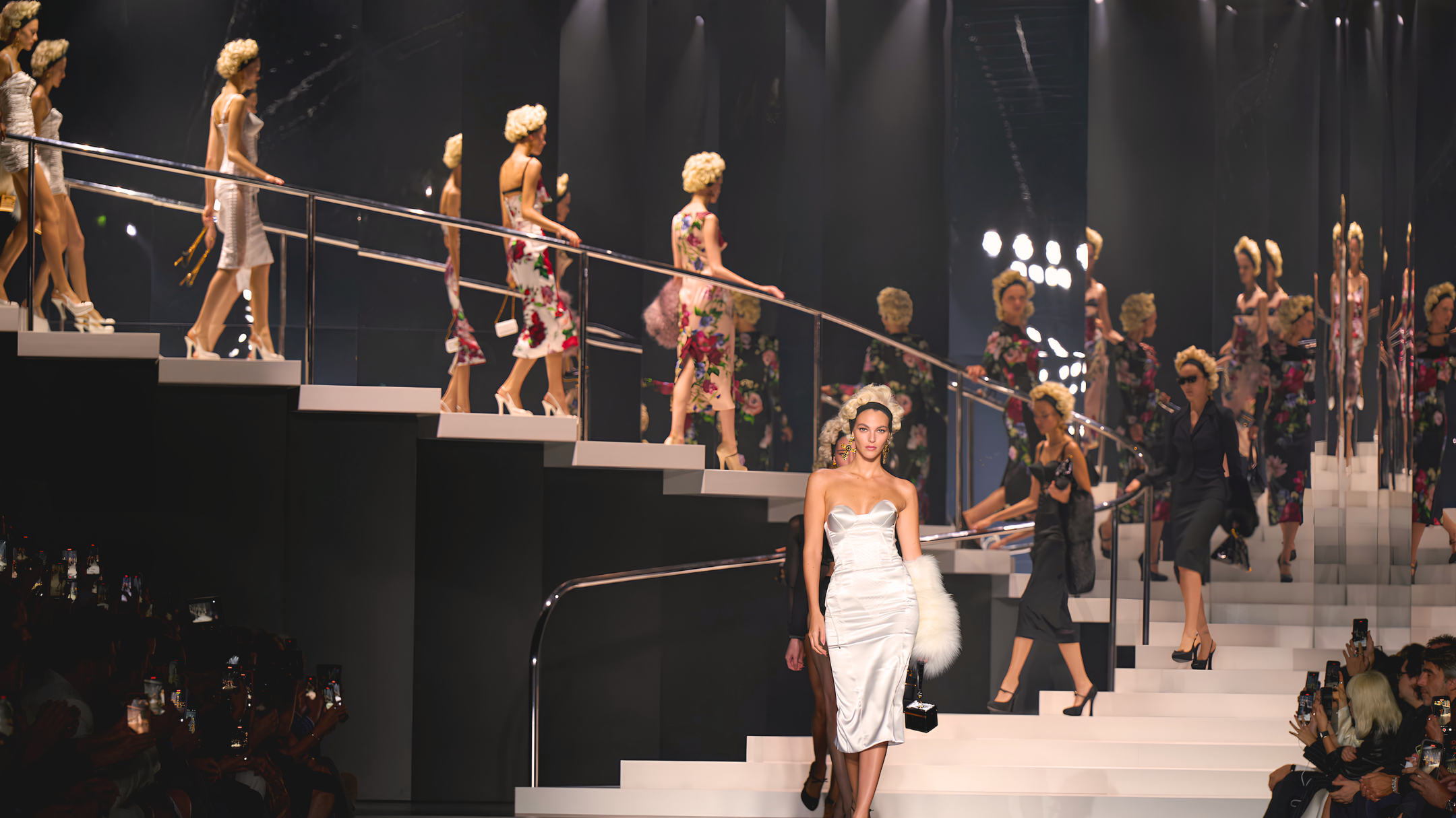 Models walk a multi-tiered runway at a luxury fashion show, showcasing elegant outfits.