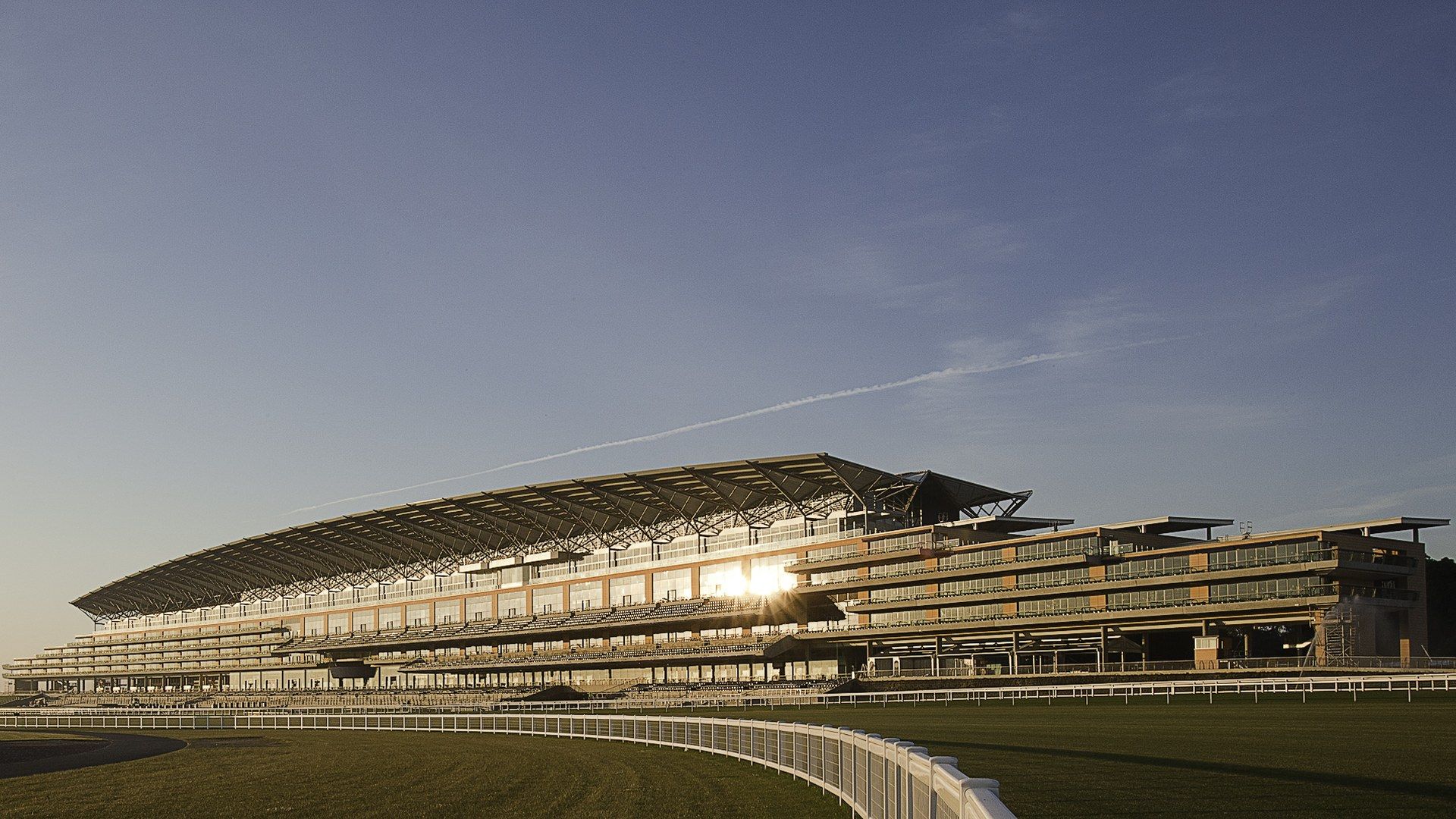 The grandstand at Ascot Racecourse in low sun, with the white running rail curving towards it