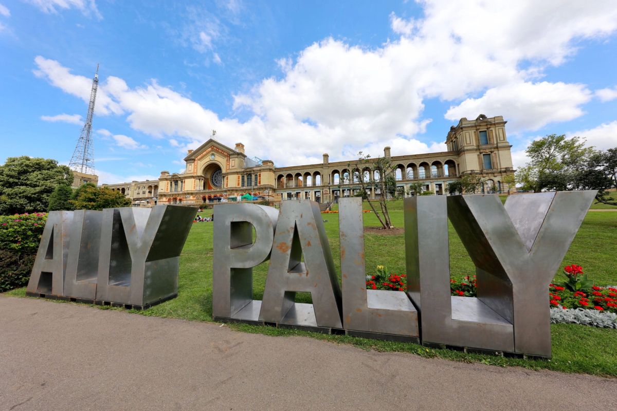 Ally Pally sign with Alexandra Palace, people relaxing on grass, under blue sky.