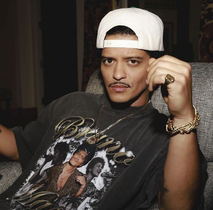 Musician Bruno Mars in a gold-script t-shirt, jewels, and cap, seated in a lounge.