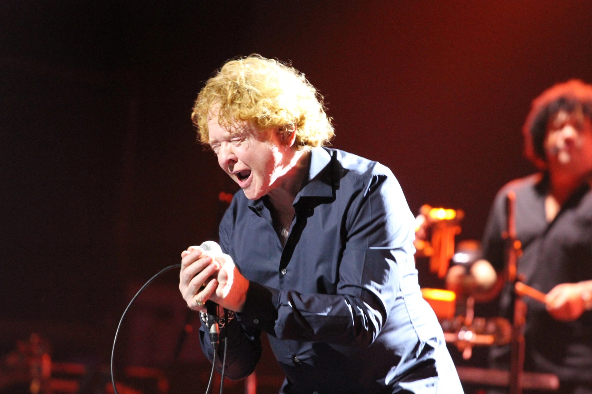 Mick Hucknall of Simply Red passionately singing on stage, eyes closed, bathed in warm light.
