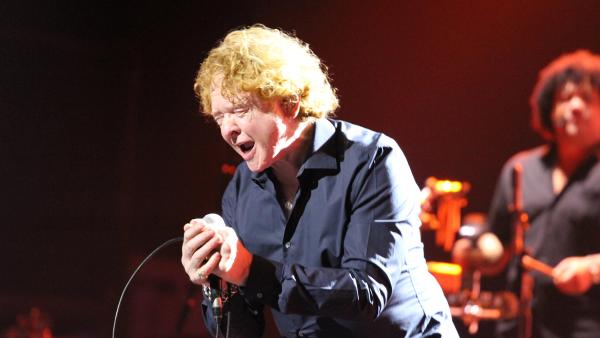 Mick Hucknall of Simply Red passionately singing on stage, eyes closed, bathed in warm light.