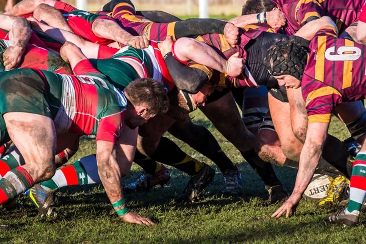 Mud-splattered players scrum for a Gilbert rugby ball on a grass pitch; number 9 visible.