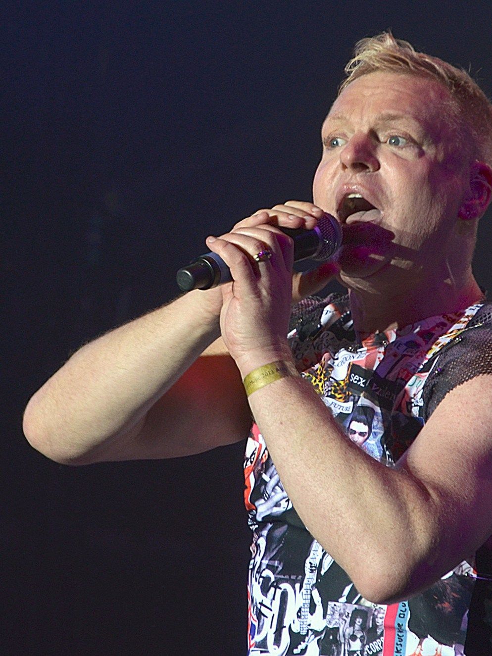 Andy Bell of Erasure singing into a microphone held in both hands, in a sleeveless printed top