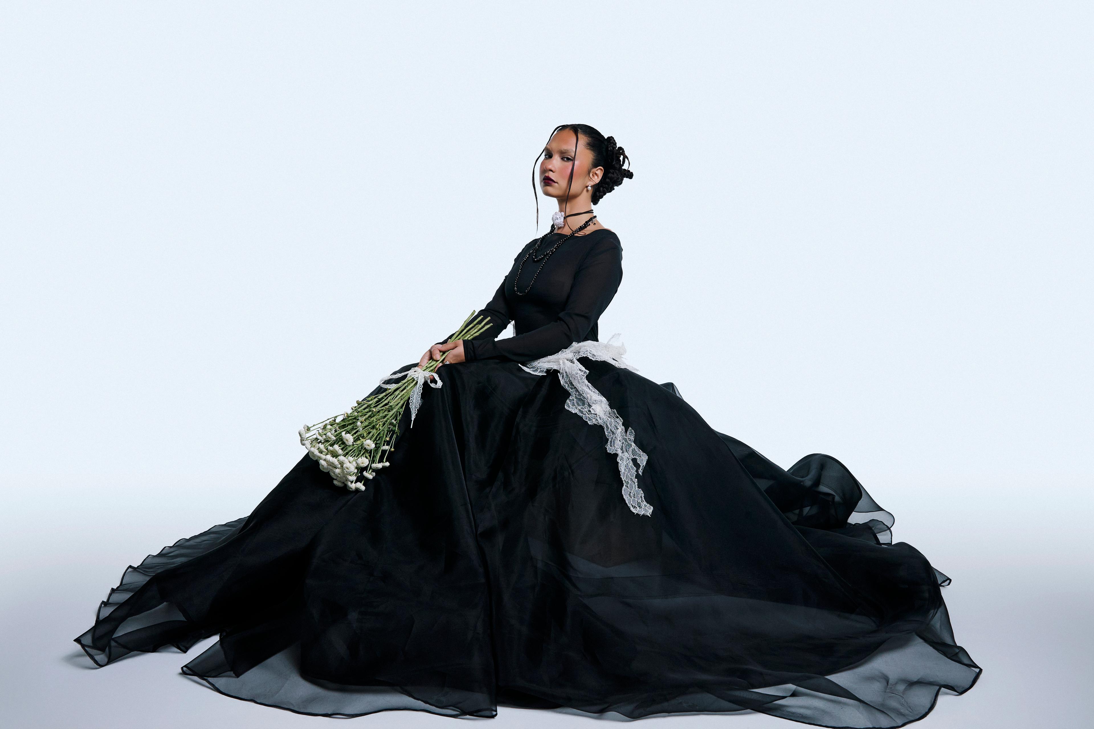 Model sitting down in a big black crinoline skirt holding white flowers.