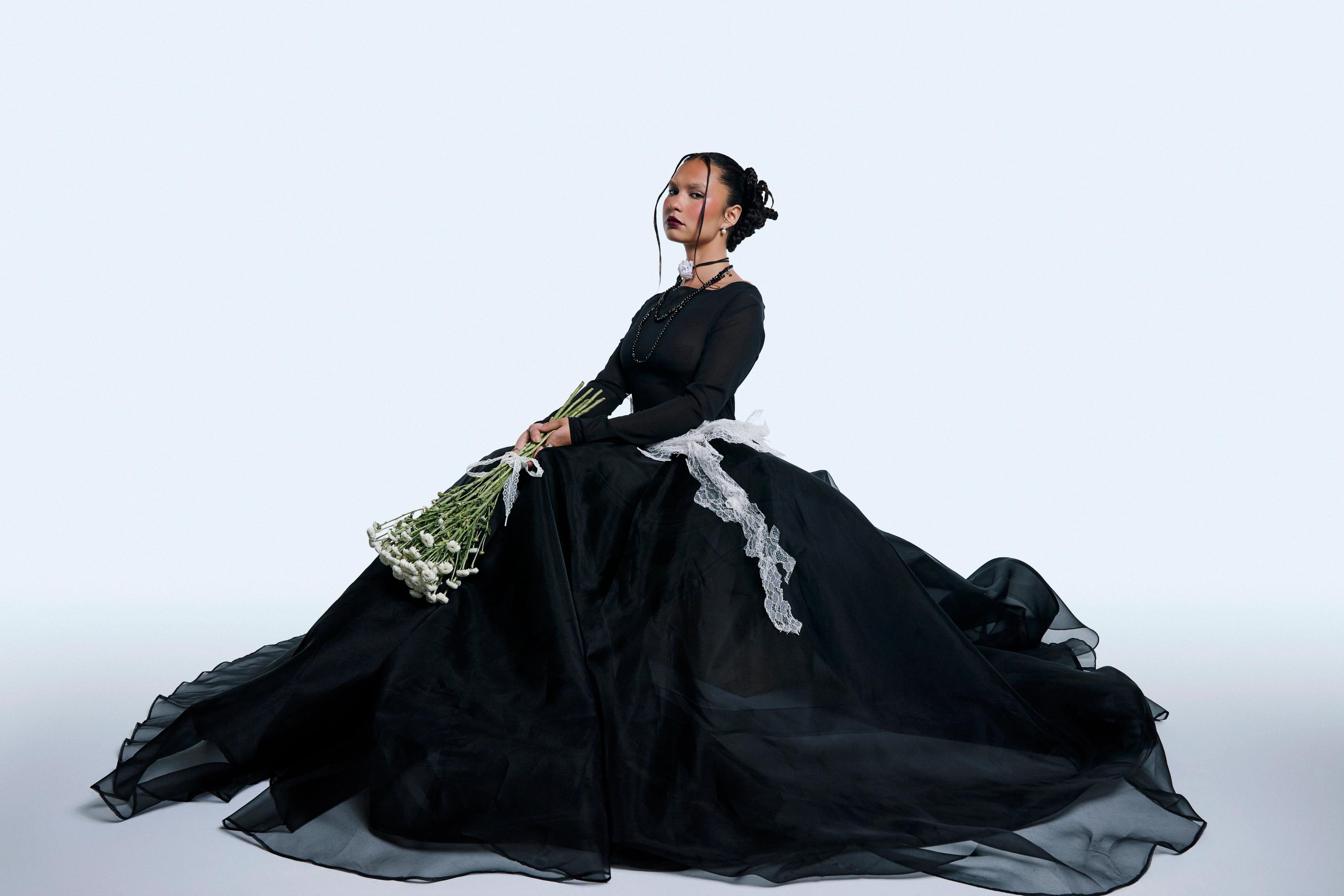 Model sitting down in a big black crinoline skirt holding white flowers.