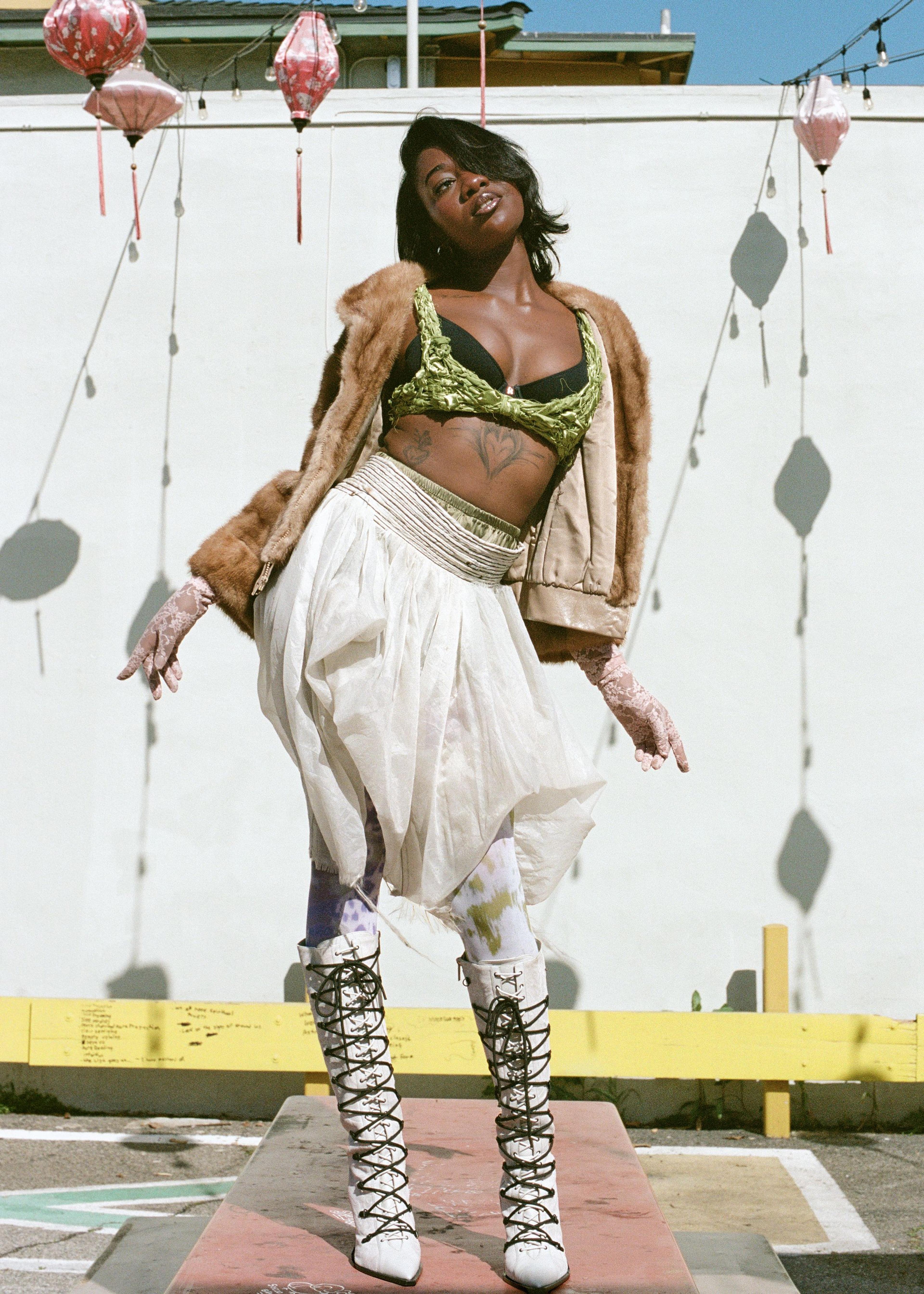 Dark skin model on a table wearing white skirt, green bra, and fur jacket in Chinatown.