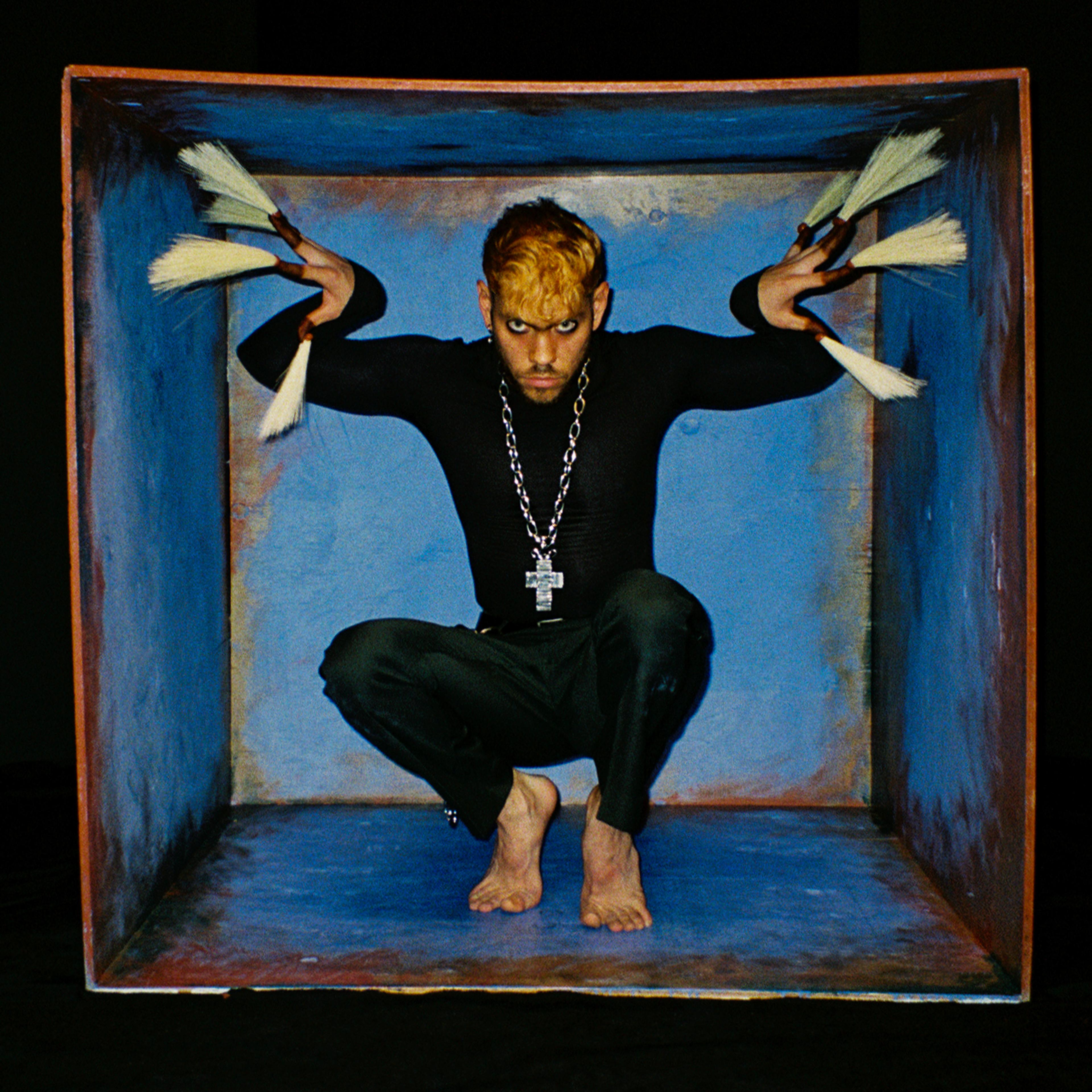 Cain Culto crouched in a blue box with white claws wearing a silver cross necklace and all black.