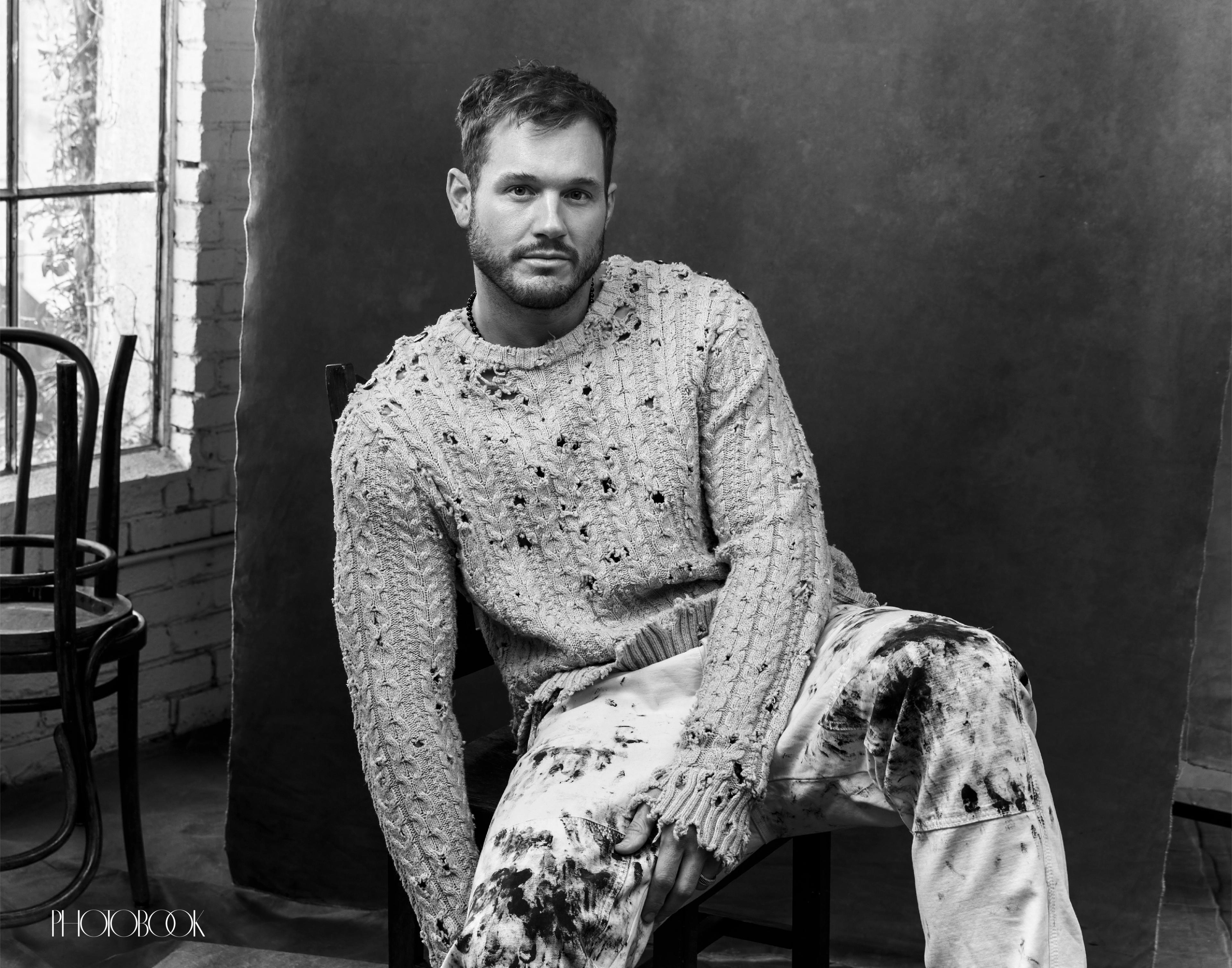 Colton Underwood wearing a tan distressed sweater facing the camera sitting down, black and white.