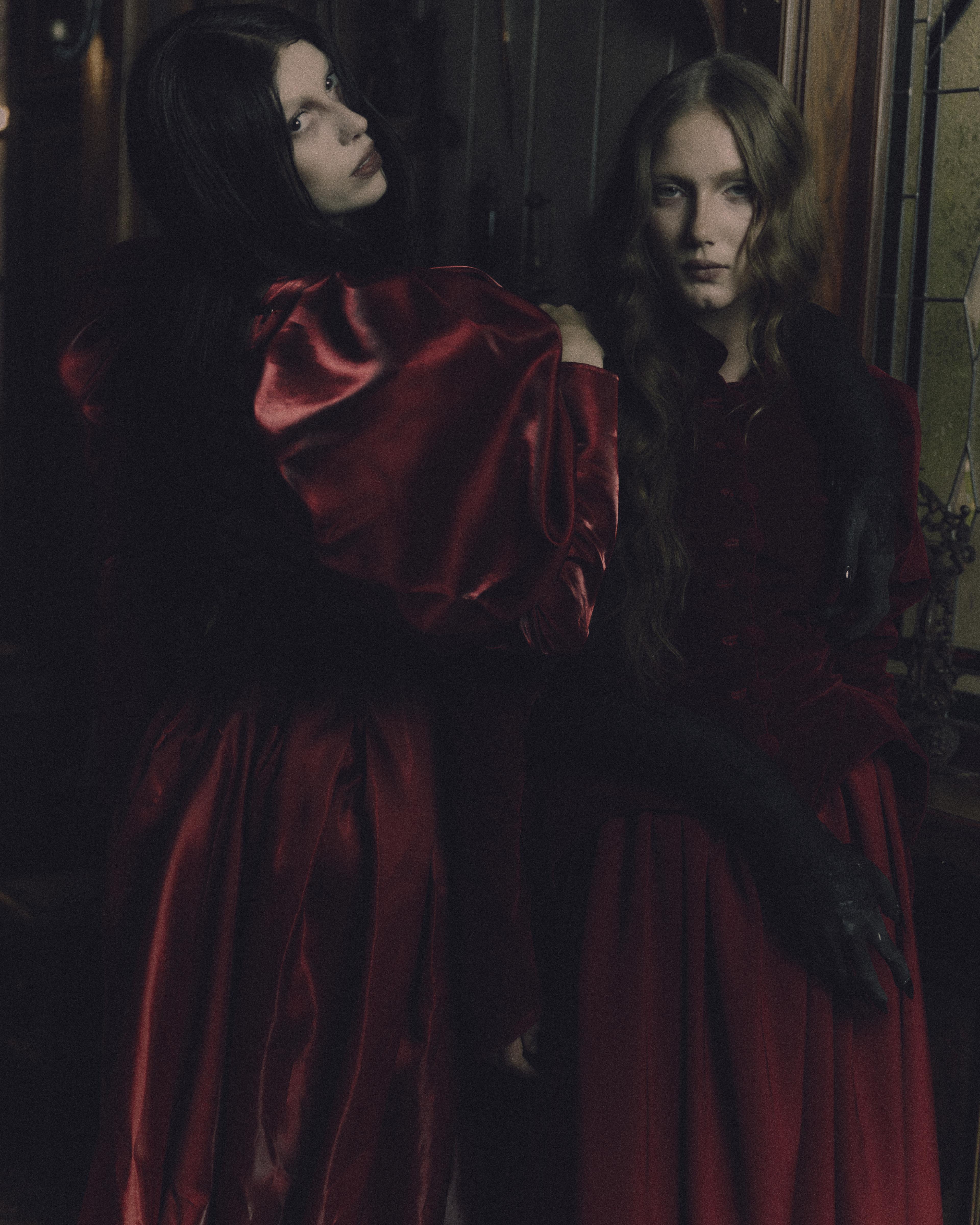 A portrait of two women wearing red custom gothic looks facing the camera.