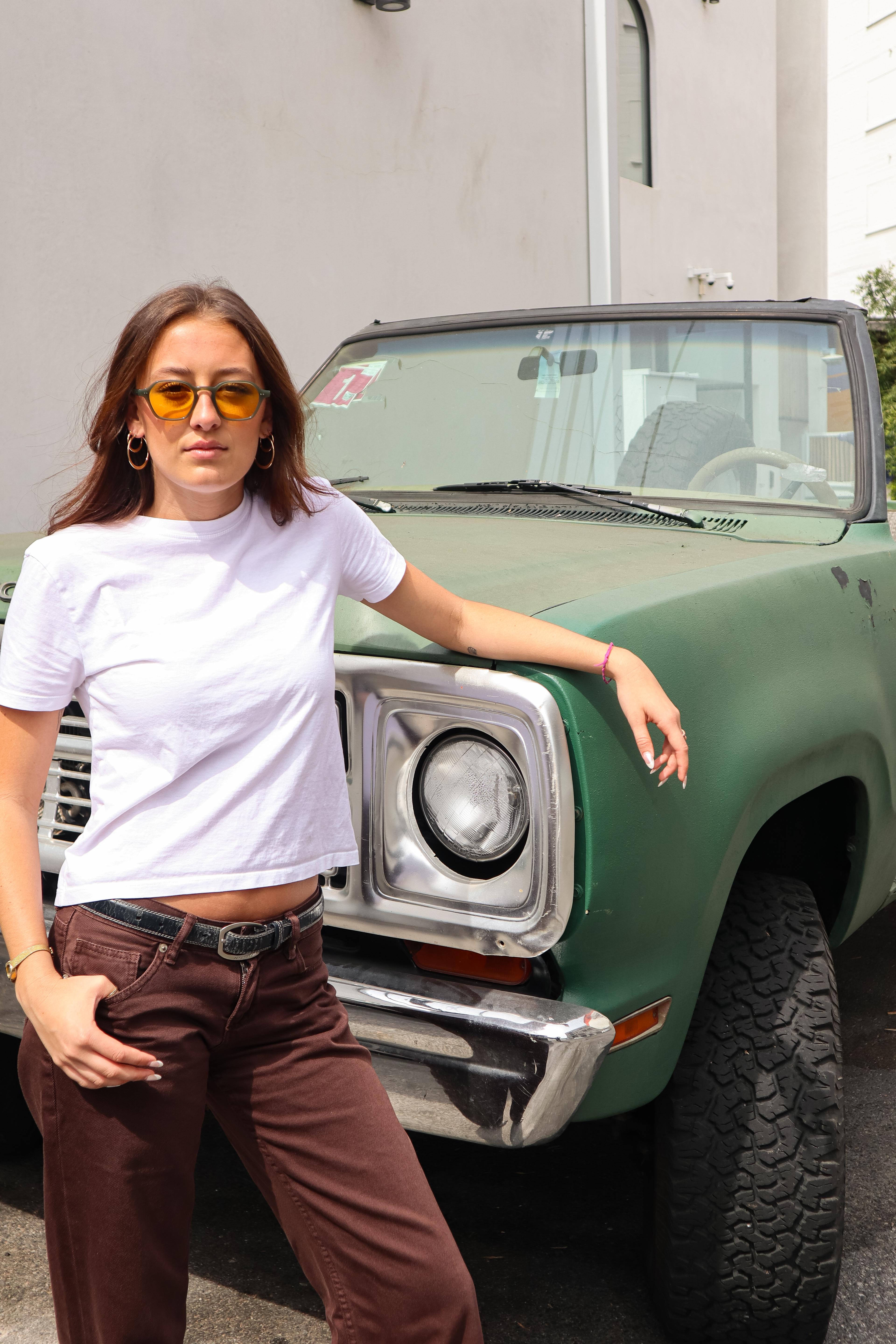 Girl with brown hair wearing yellow glasses in front of a green old Bronco with white tshirt and brown pants.