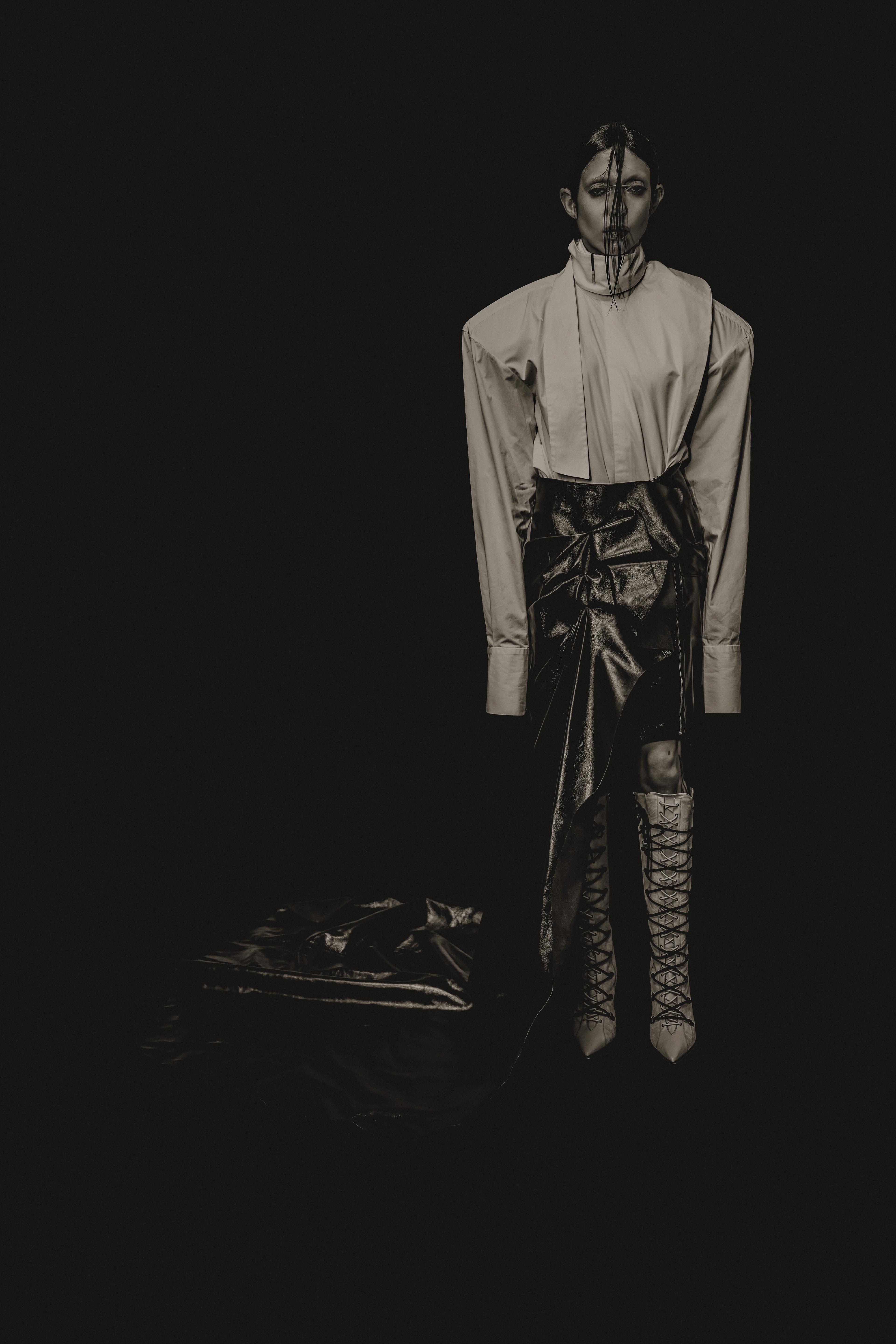 DeathByRomy wearing a big jacket and white boots in black and white standing straight.