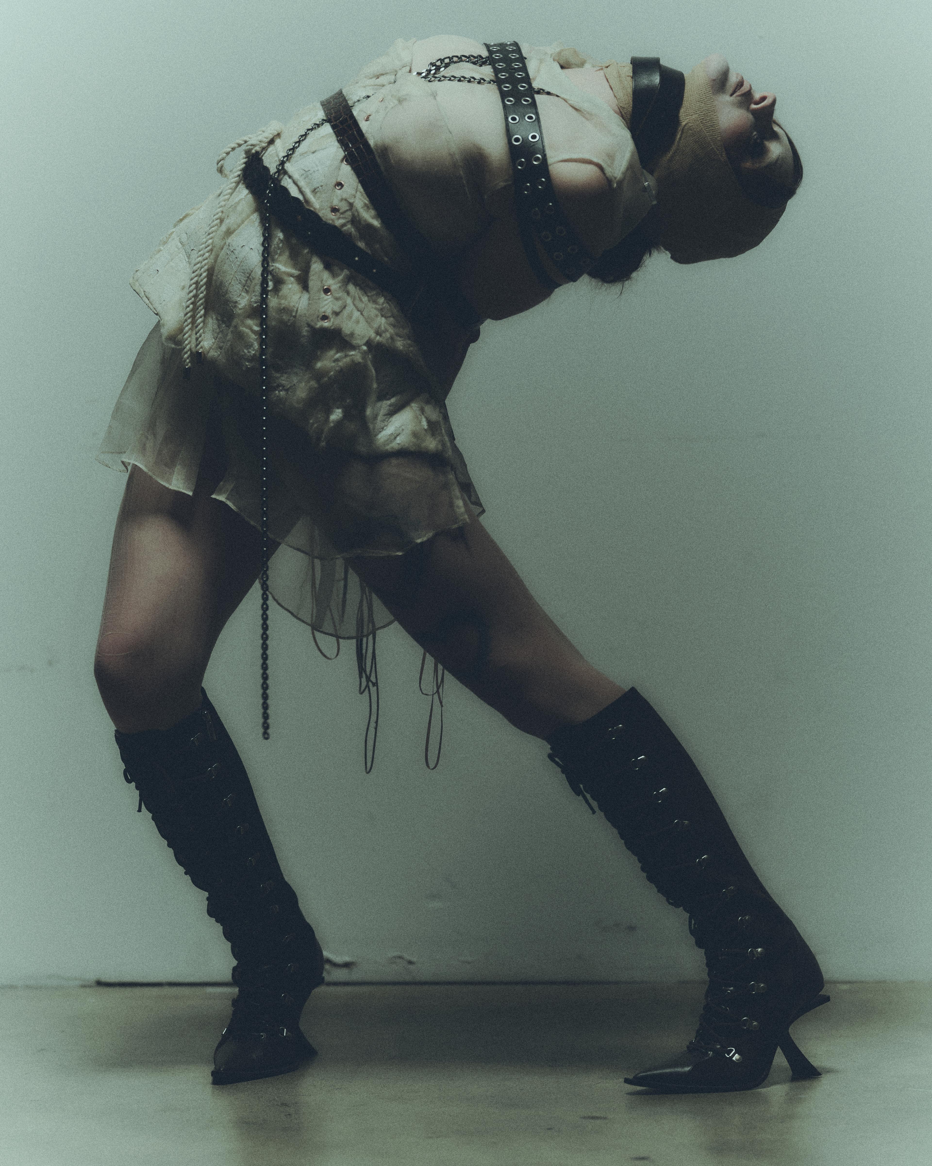 Girl with head bandages wearing cream layered look strapped in with belts and brown boots bent over.