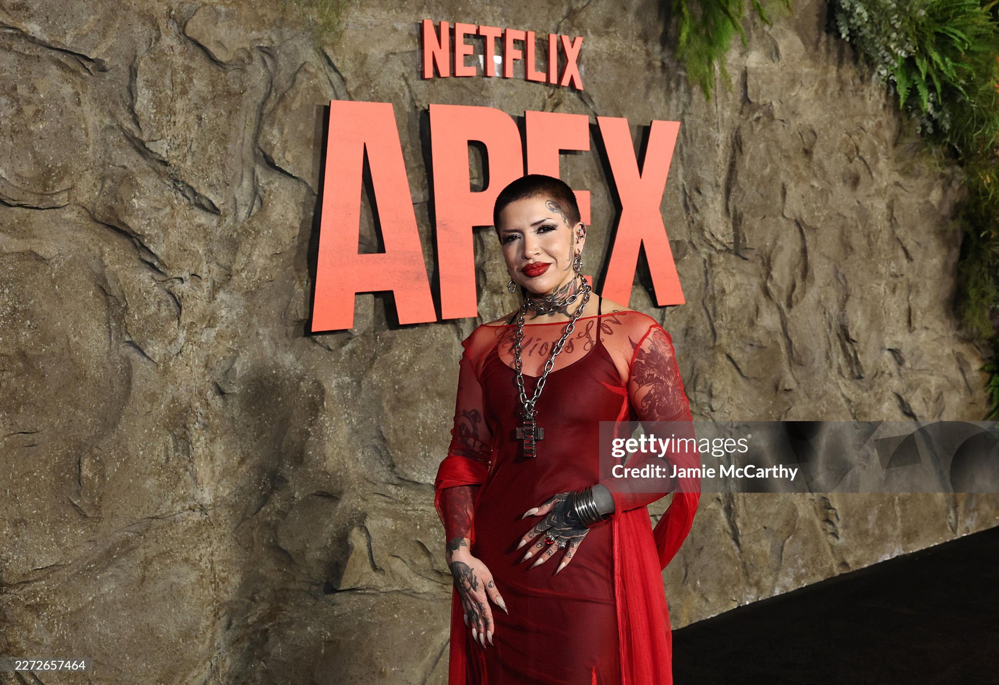 Nina Pool in a red sheer gown on a red carpet movie premier with silver cross necklace.