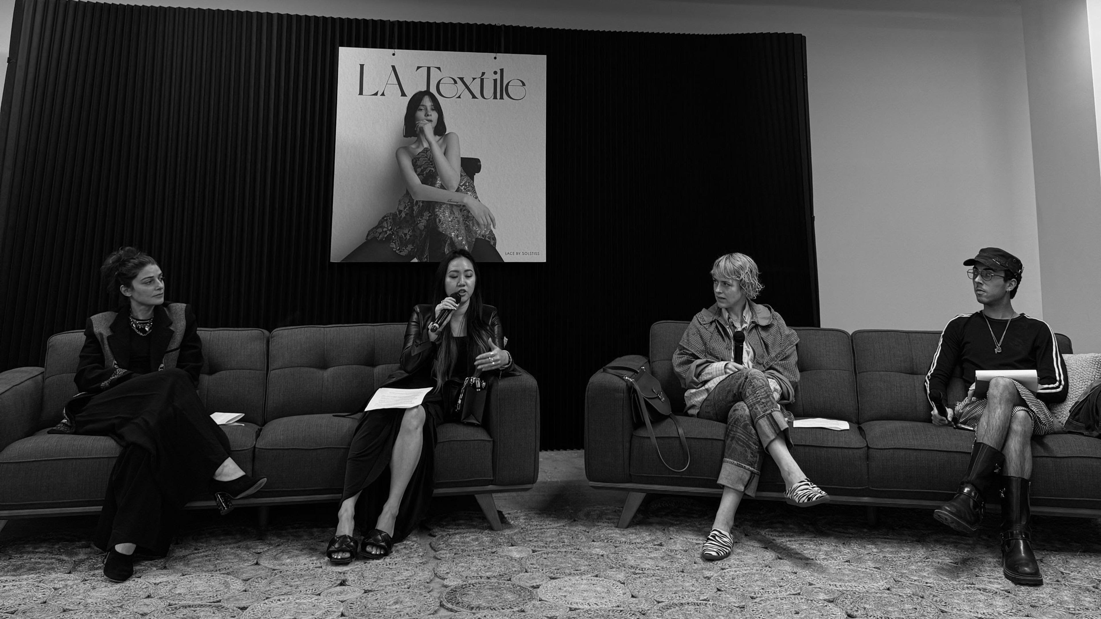 Black and white photo of four people sitting on couches with microphones doing a panel talk.