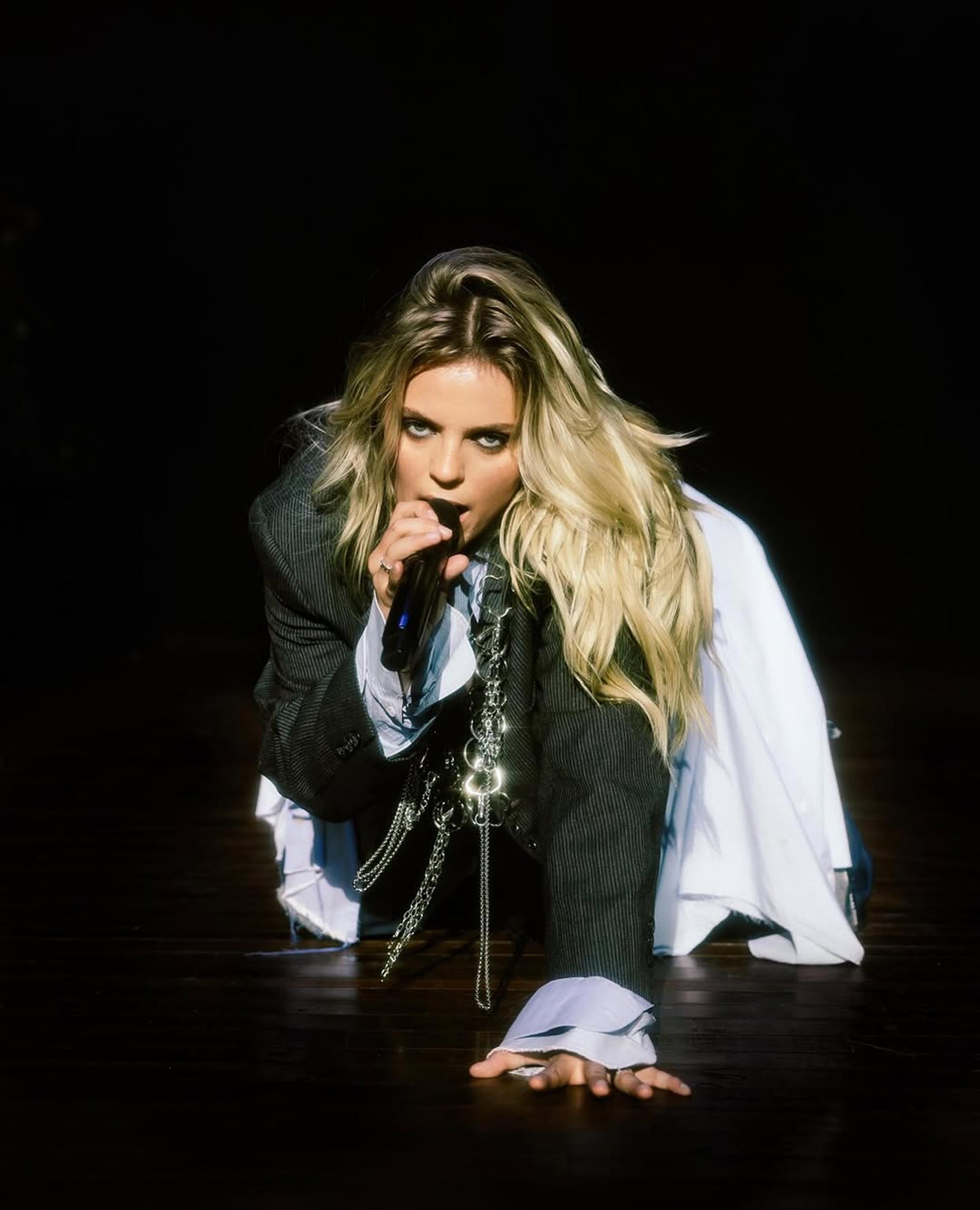 Renee Rapp crawling to the camera with microphone in suiting.
