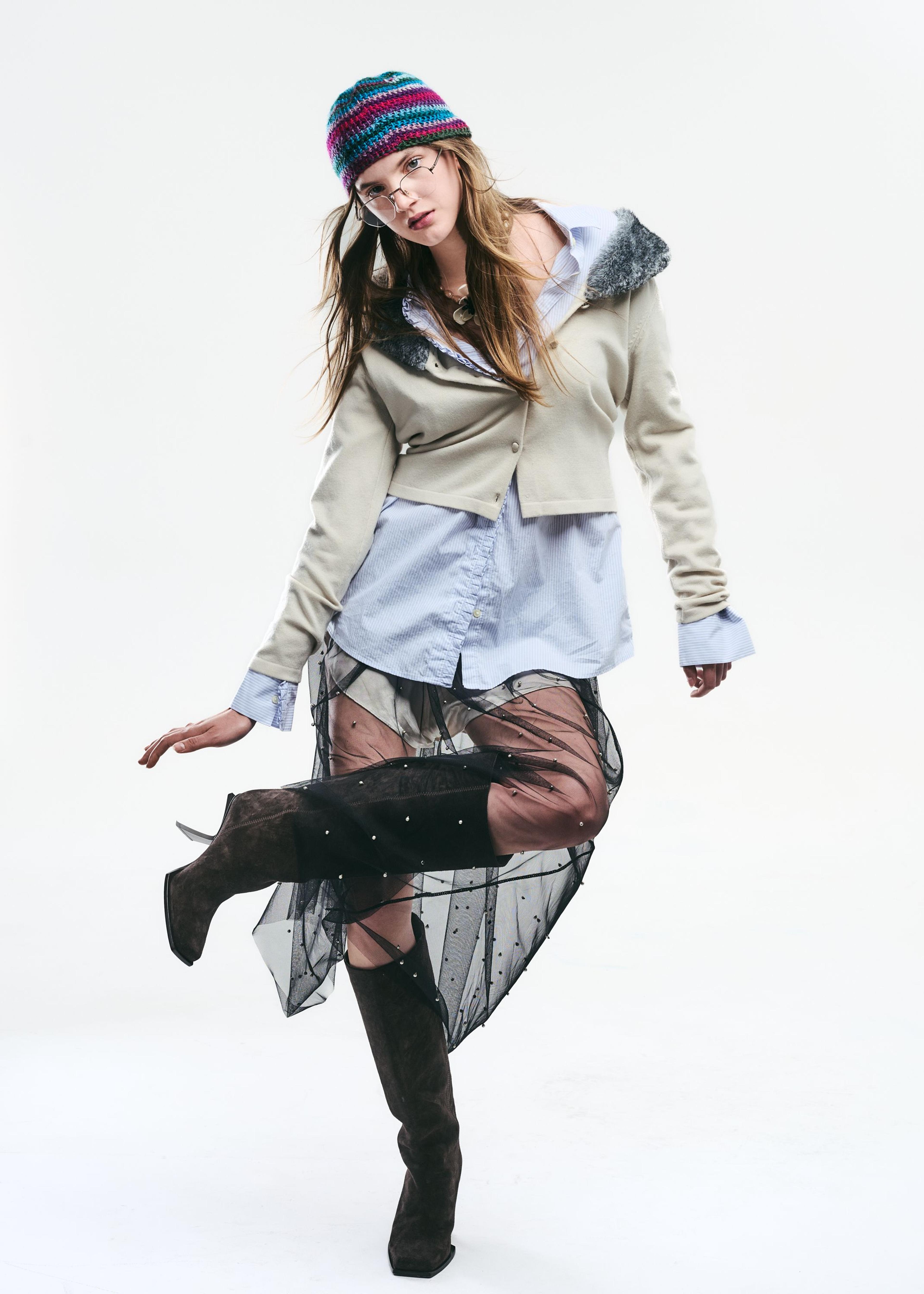 Blonde model with glasses and beanie wearing a cardigan, shirt, black sheer skirt, and boots standing on one foot.