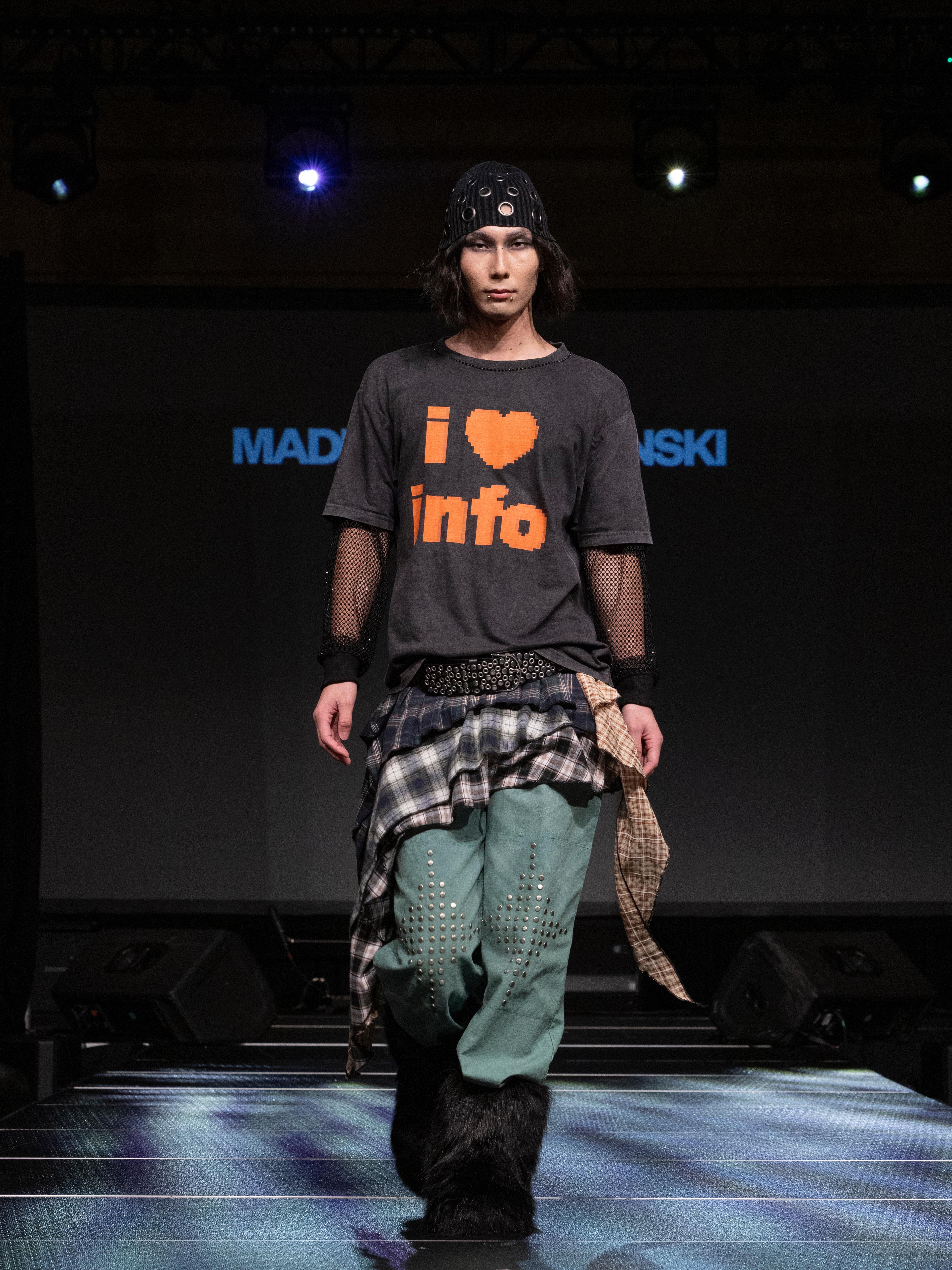 Model in grey beanie with graphic tee and green pants walking a runway.