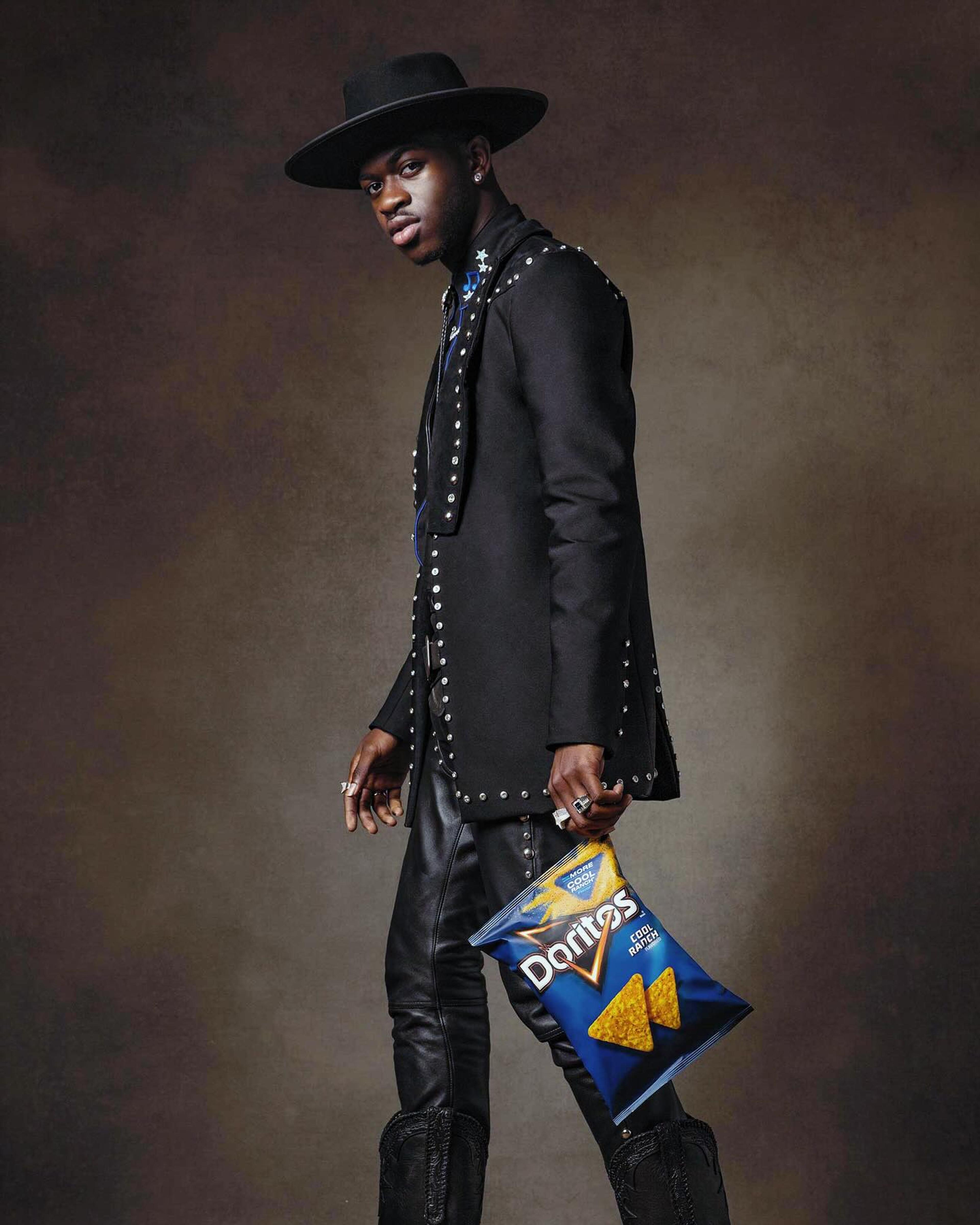Lil Nas X in a studded suit all black holding a dorritos bag.