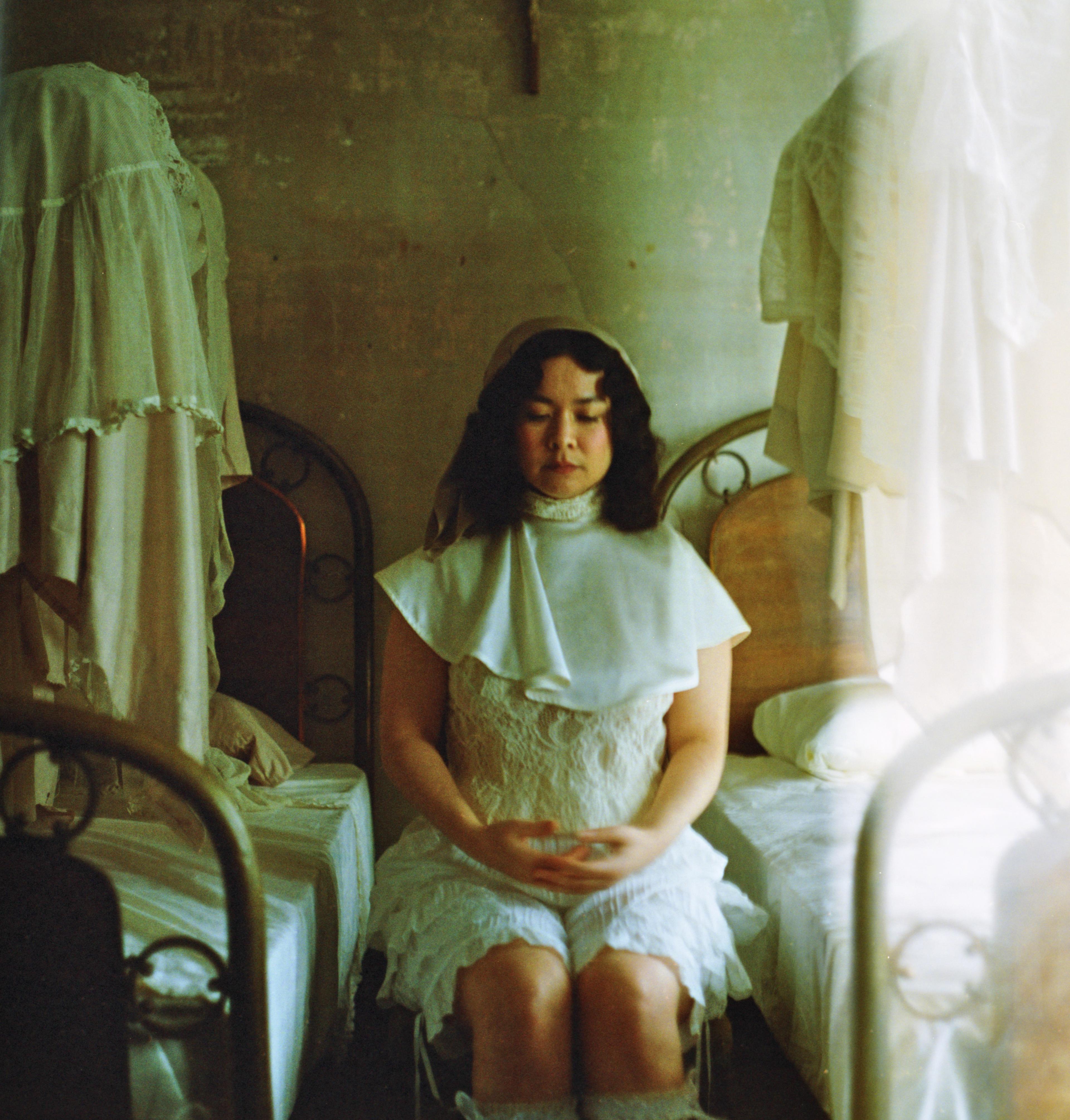 Mitski sitting in a vintage bed wearing all white with her eyes closed looking up.