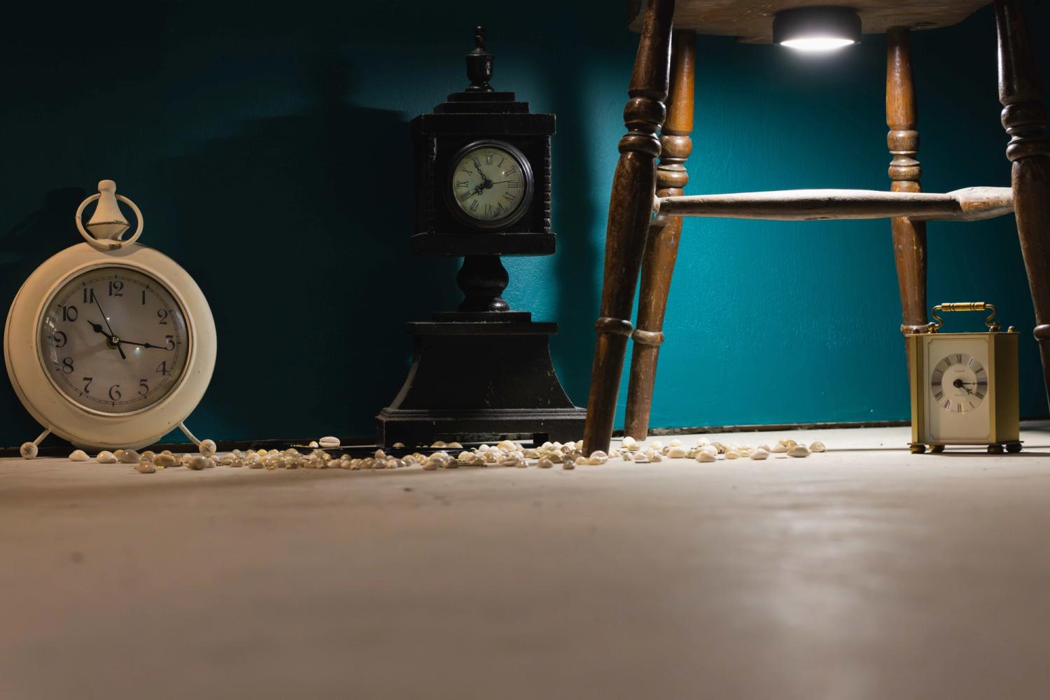 A low-angle scene showing three vintage clocks beneath a wooden chair lit by a small overhead light, with scattered shells or beads across the floor against a wall