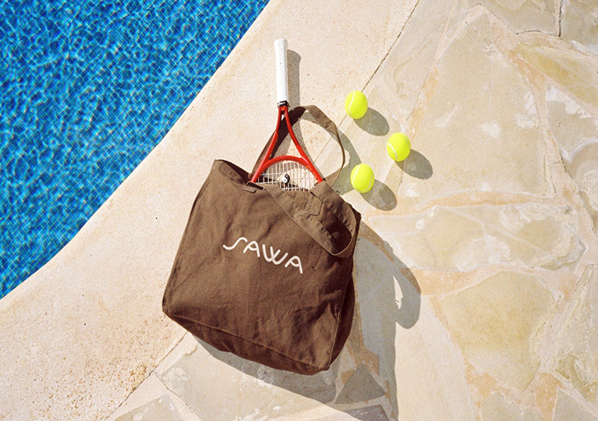 A SAWA tote bag with a tennis racket and balls beside a pool