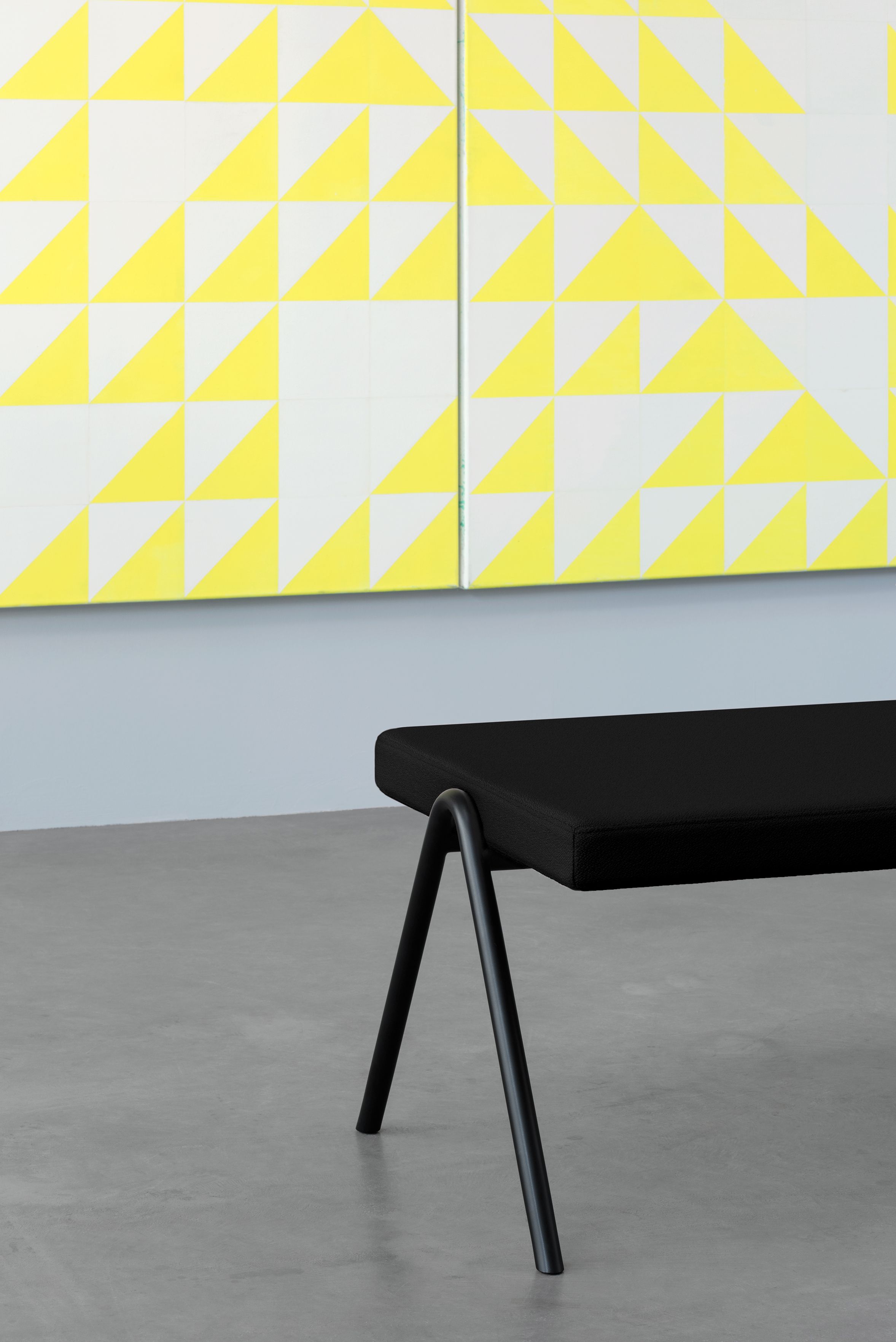 a black seating in front of a yellow artwork, grey floor and wall