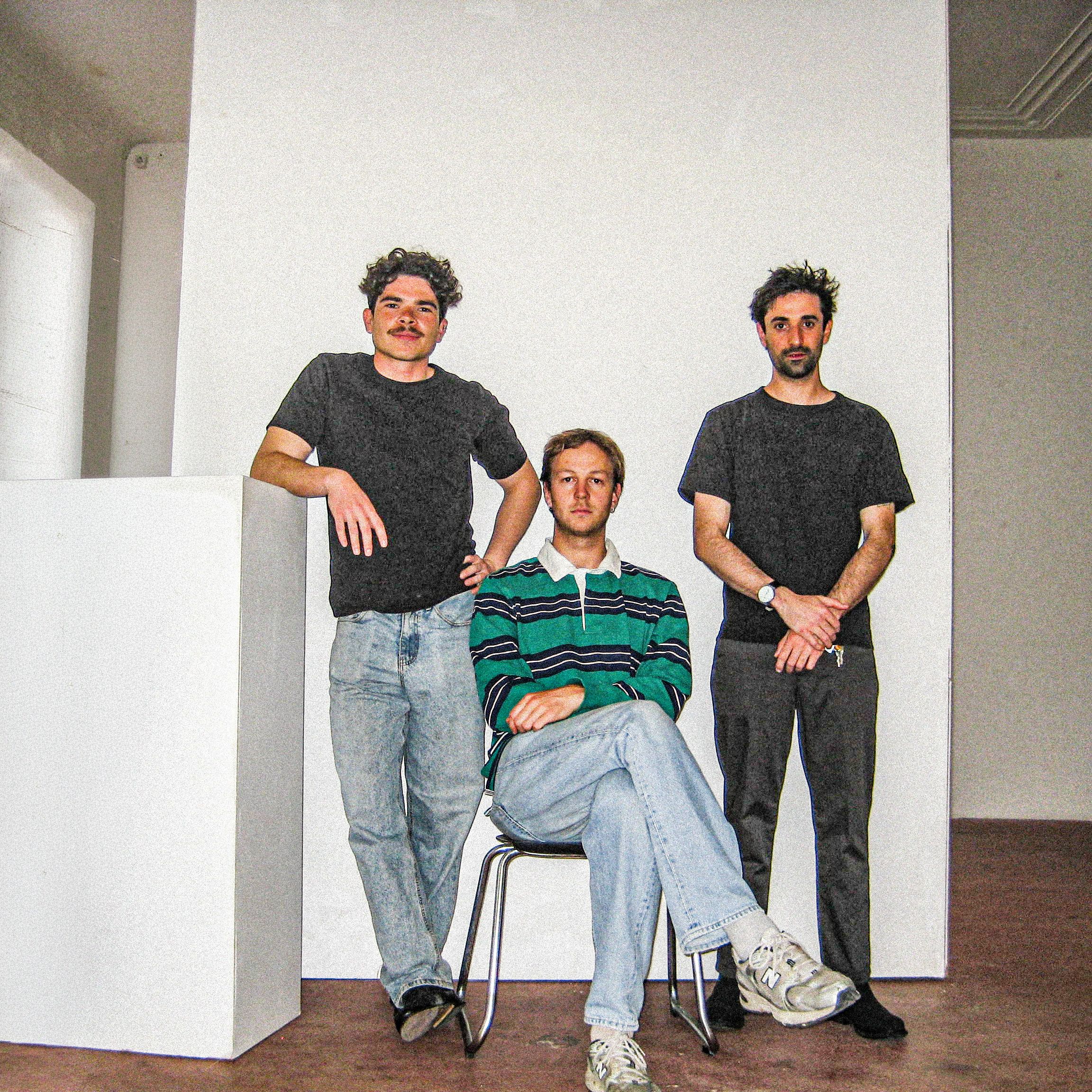 Three men in front of a white wall, one is sitting on a chair