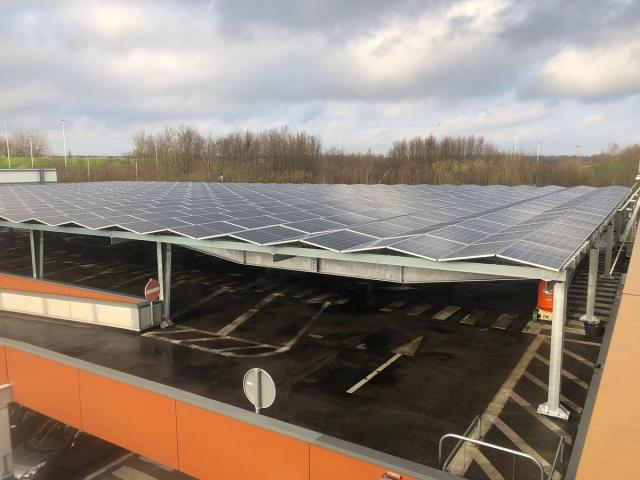 Solar-panel carport installation covering a large parking area at a Decathlon site. Rows of photovoltaic panels are mounted on steel canopy structures above parking spaces, providing both renewable energy generation and vehicle shelter. The scene is viewed from an elevated position under a cloudy sky, with trees visible in the background.