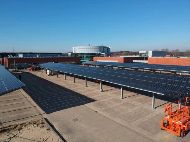 Large solar-panel carport installation in front of a modern commercial or institutional building. Multiple rows of photovoltaic panels are mounted on elevated steel structures above a paved parking area, with construction equipment visible on site under a clear blue sky.