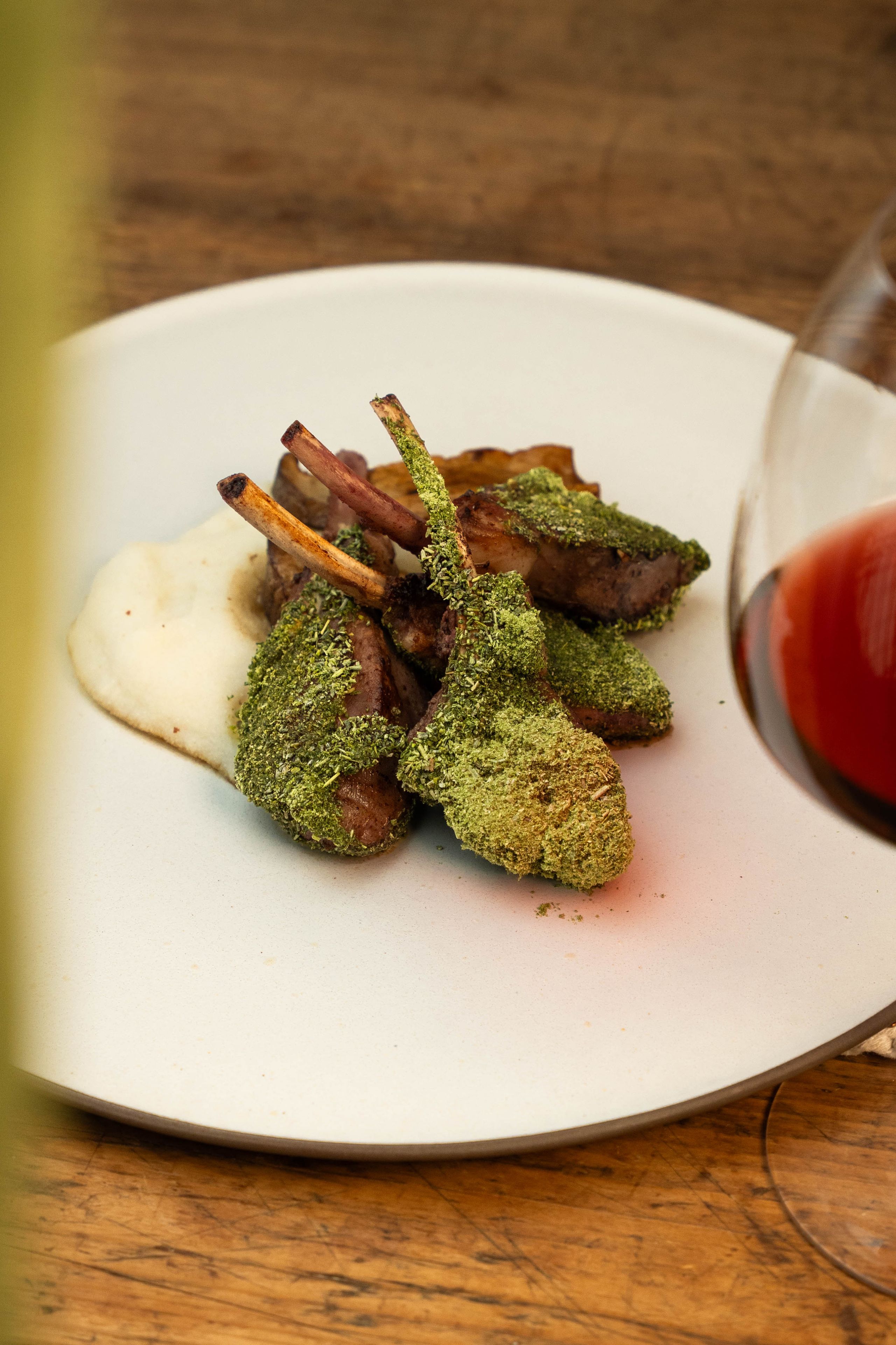 Costolette di agnello con crumble verde su crema bianca, su piatto bianco con bicchiere di vino rosso sullo sfondo