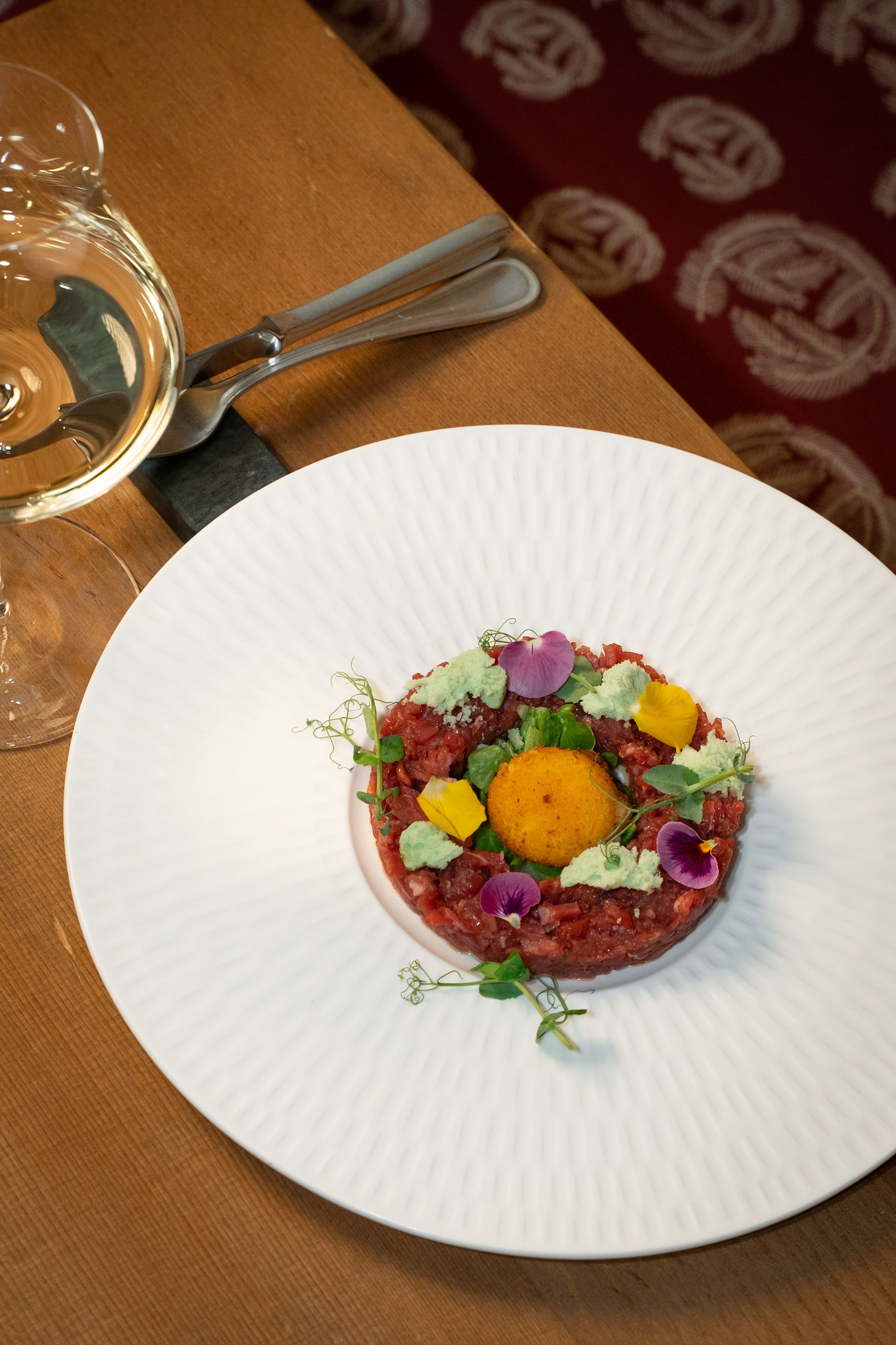 Tartare di carne con tuorlo d'uovo, crema verde e fiori eduli in piatto bianco su tavolo di legno