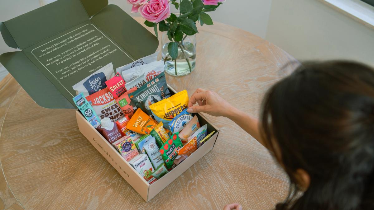 Person selecting a packaged snack from an open The Snack Edit school snack box