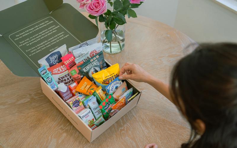 Person selecting a packaged snack from an open The Snack Edit school snack box