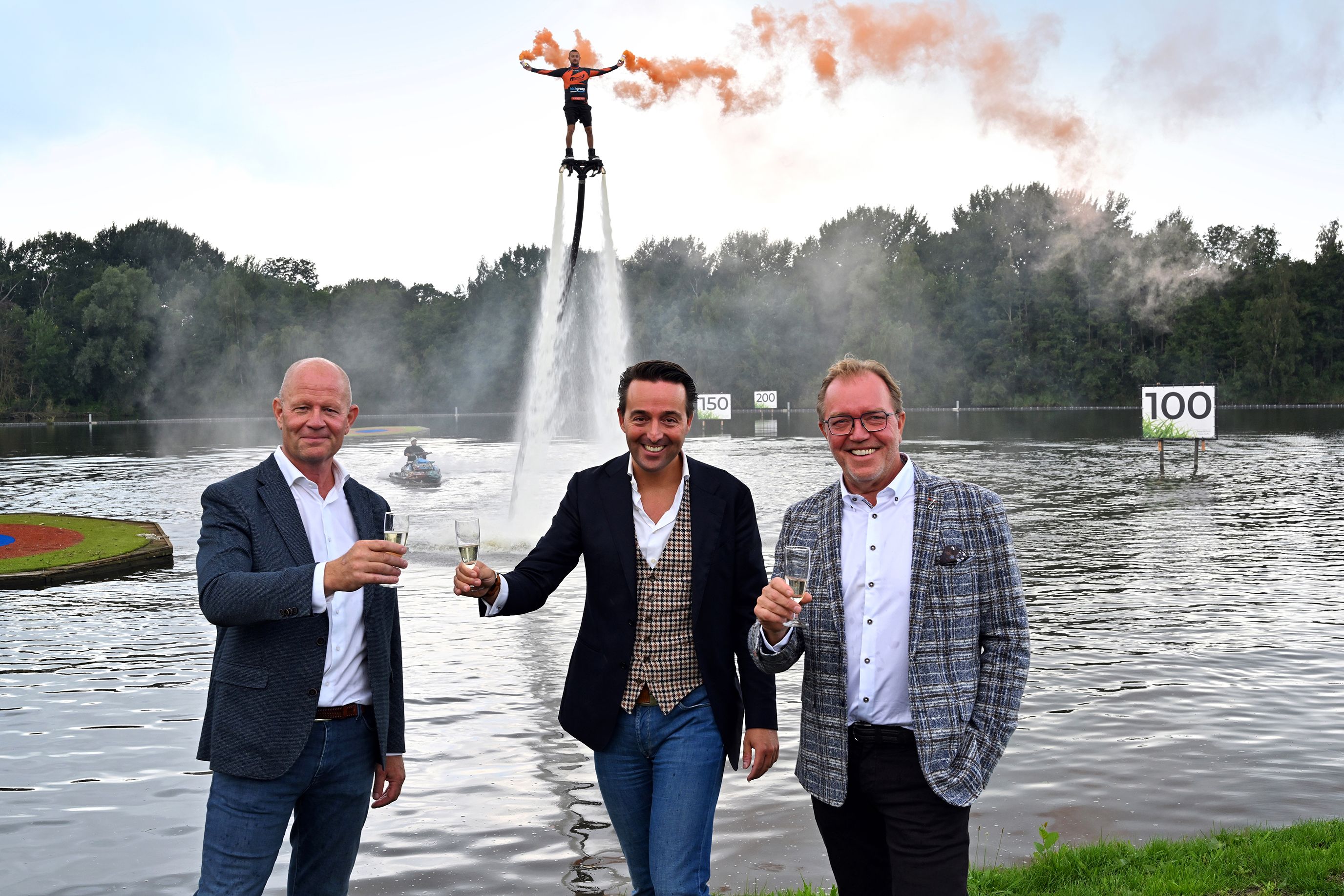 Hans Schaap, Mark Marshall en Joris Slooten brengen een toost uit op de heropening van Golfpark Naarderbos, terwijl een flyboarder met oranje rookfakkels over het water vliegt