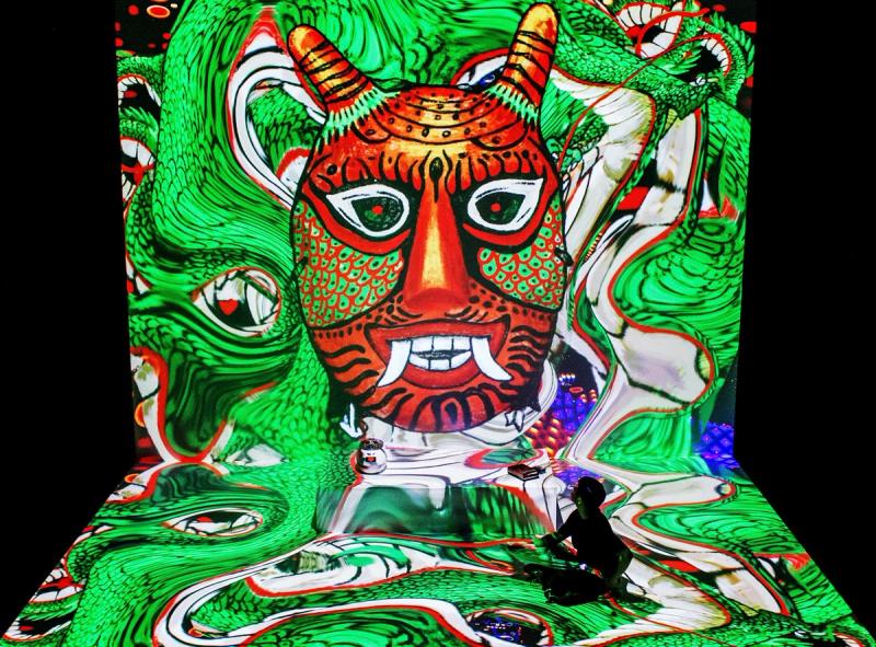 Man sitting on the floor with a big projection of a devils face and lots of colours on the floor an wall