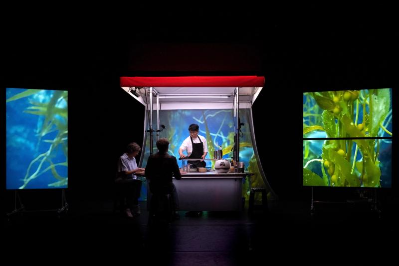 Man cooking and serving two women from a food stand. Projections of plants on both sides