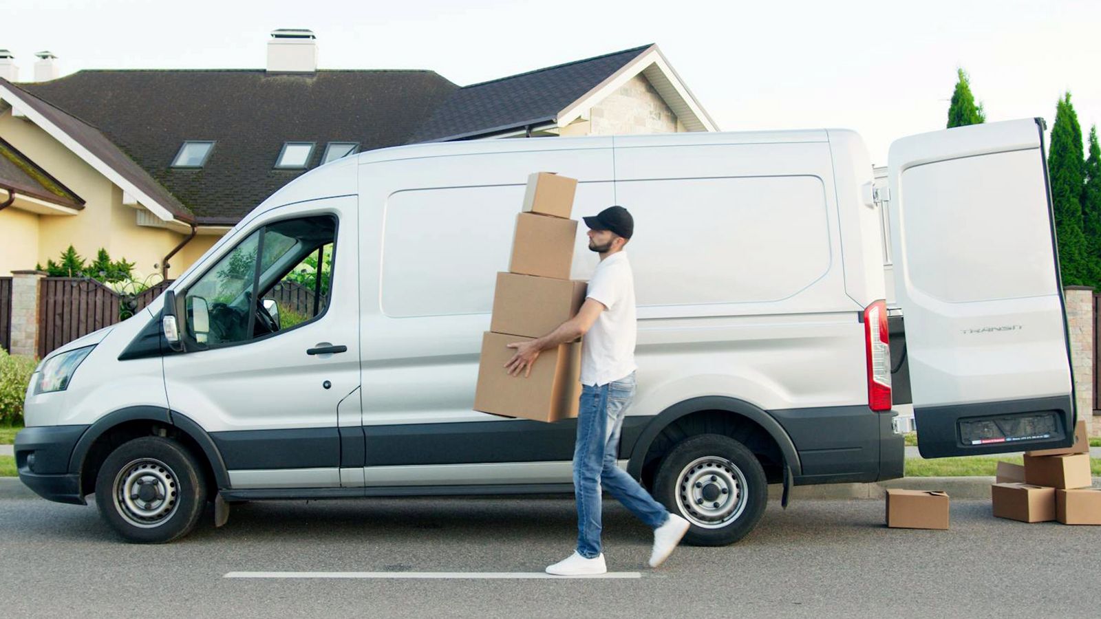A field worker carries stacked cardboard boxes to a white service van parked outside a suburban home.