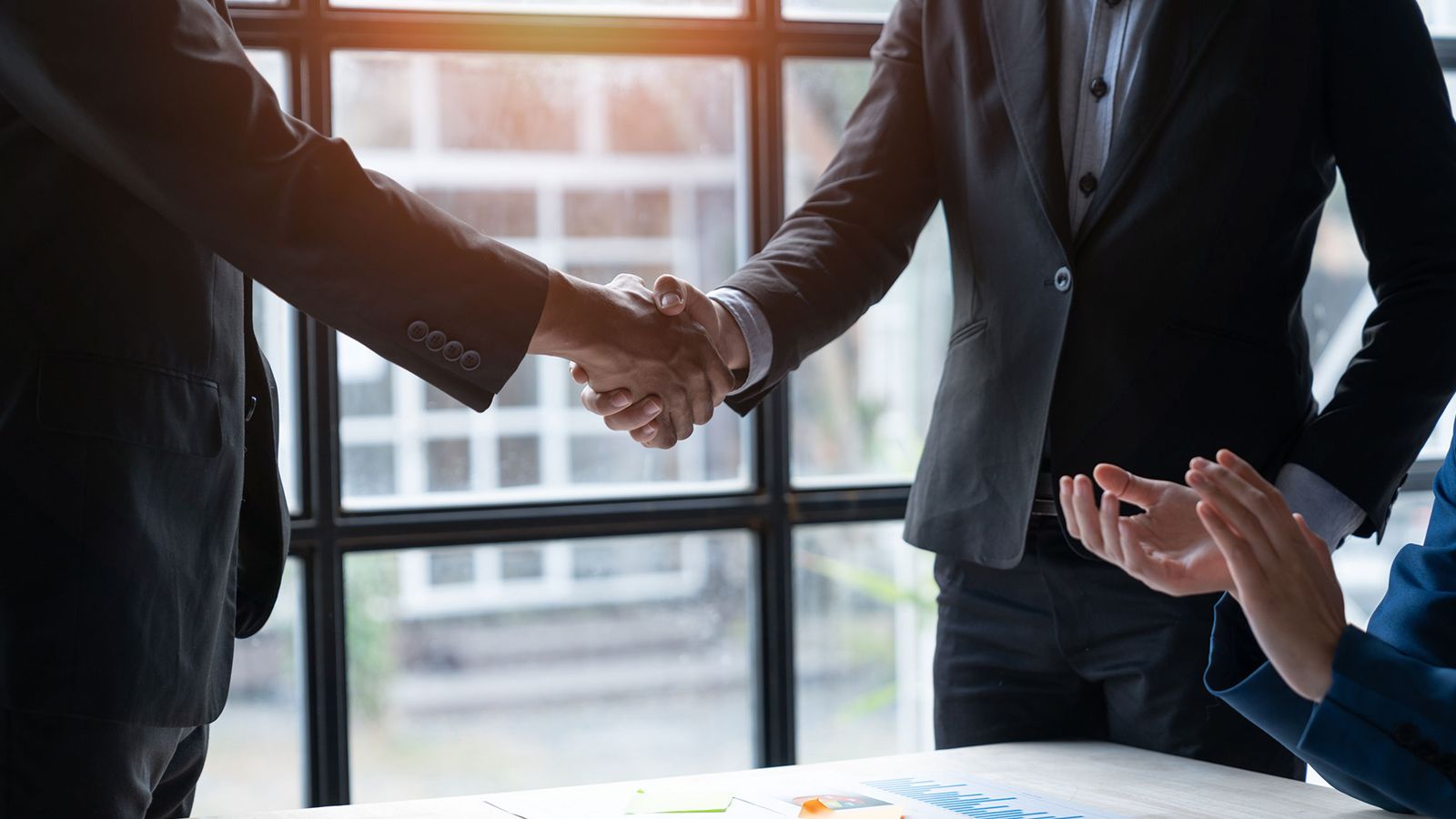 Two professionals shaking hands across a desk in a sunlit office, with a printed performance chart on the table.