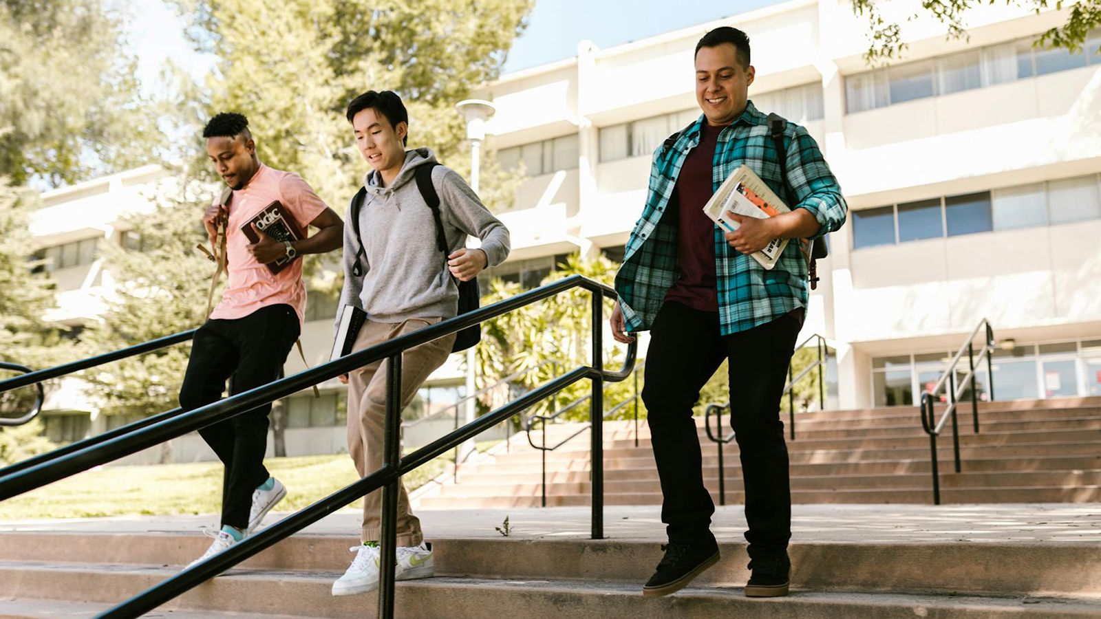 Three college students carrying books walk down exterior campus stairs on a sunny day.