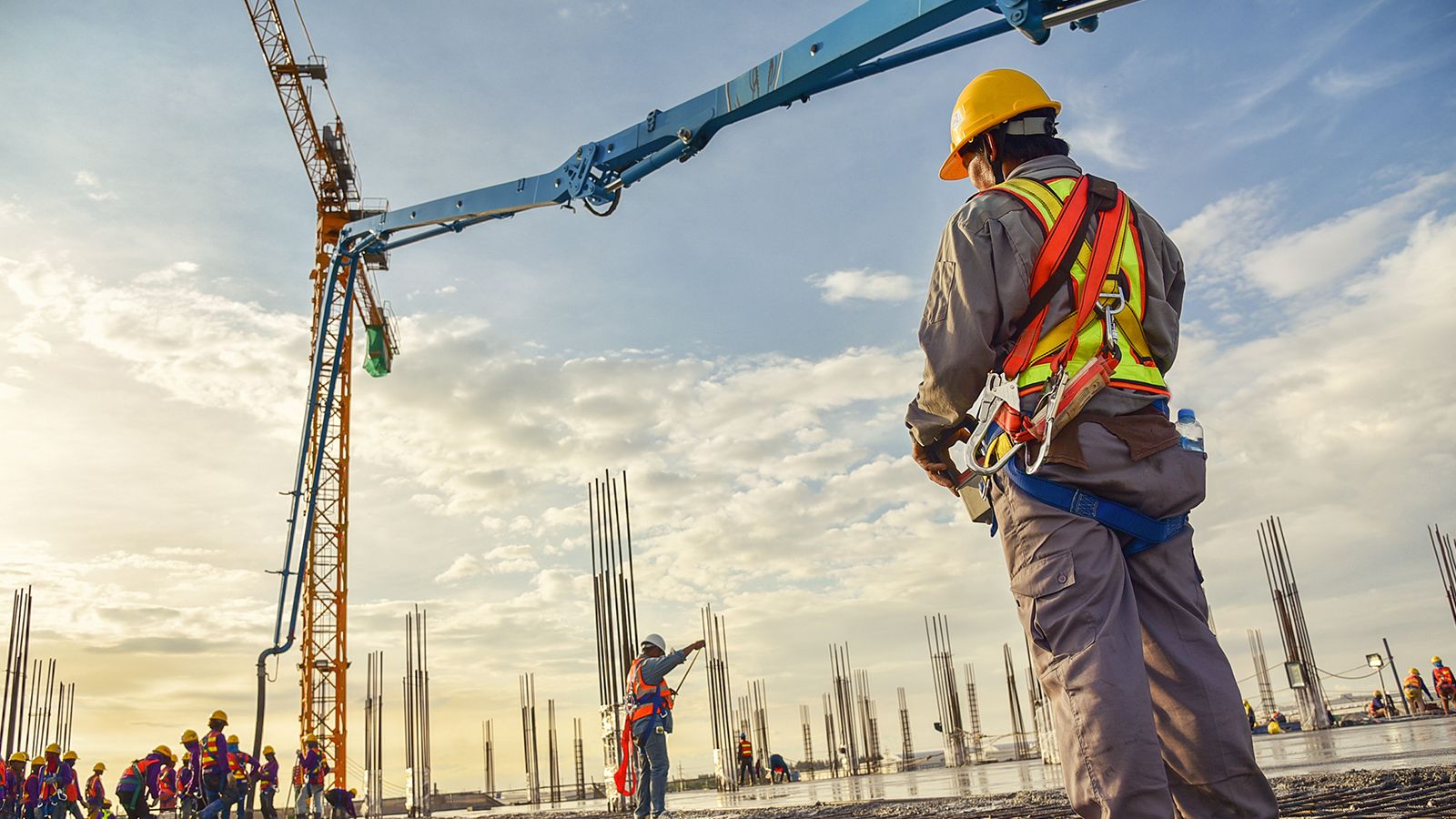 A construction worker in a safety harness and hard hat watches a concrete pump boom over a building foundation at sunrise.