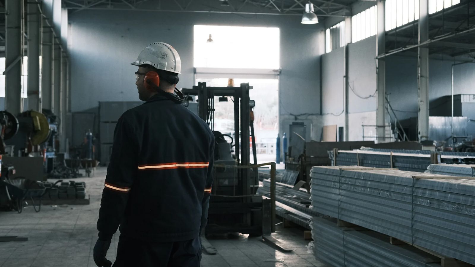 A worker in a hard hat and high-visibility jacket walks through an industrial plant past a forklift and stacked metal stock.