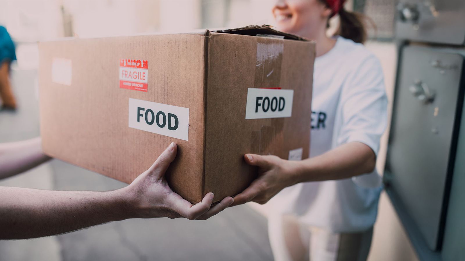 A volunteer in a branded shirt hands a cardboard box marked FOOD to another person at a community donation drive.