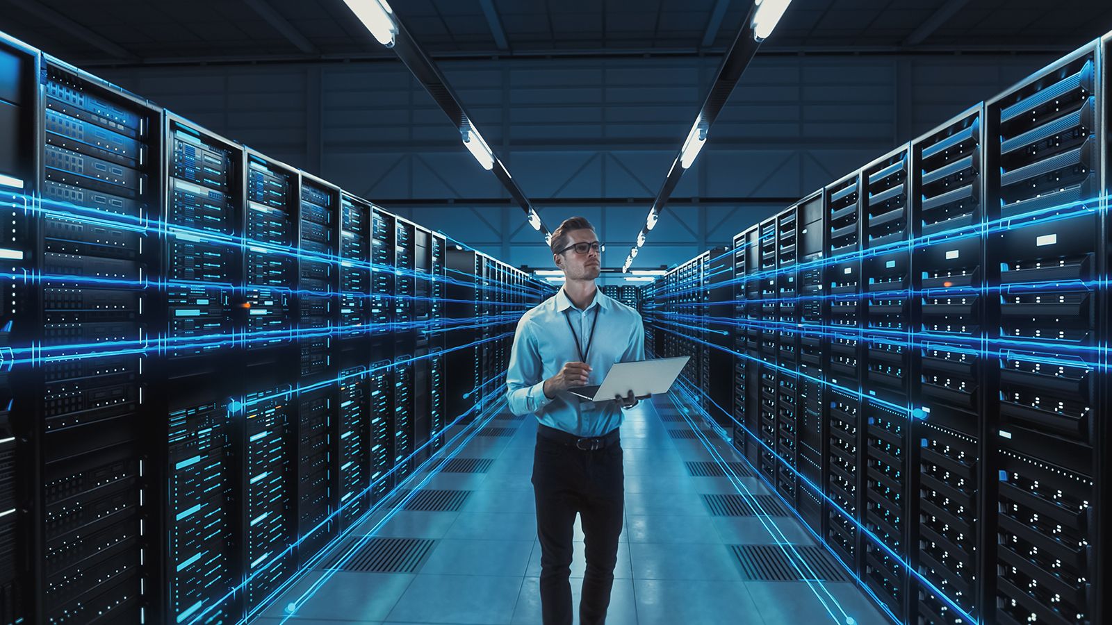 A technician holding a laptop walks down the aisle of a data center between rows of servers lit with blue light.