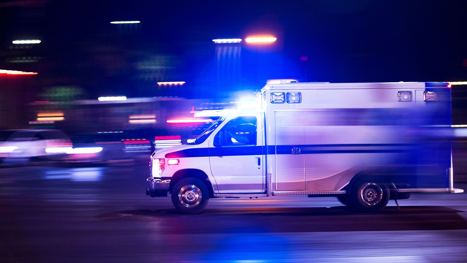 A white-and-blue ambulance speeds along a city street at night, its emergency lights streaking against the dark.
