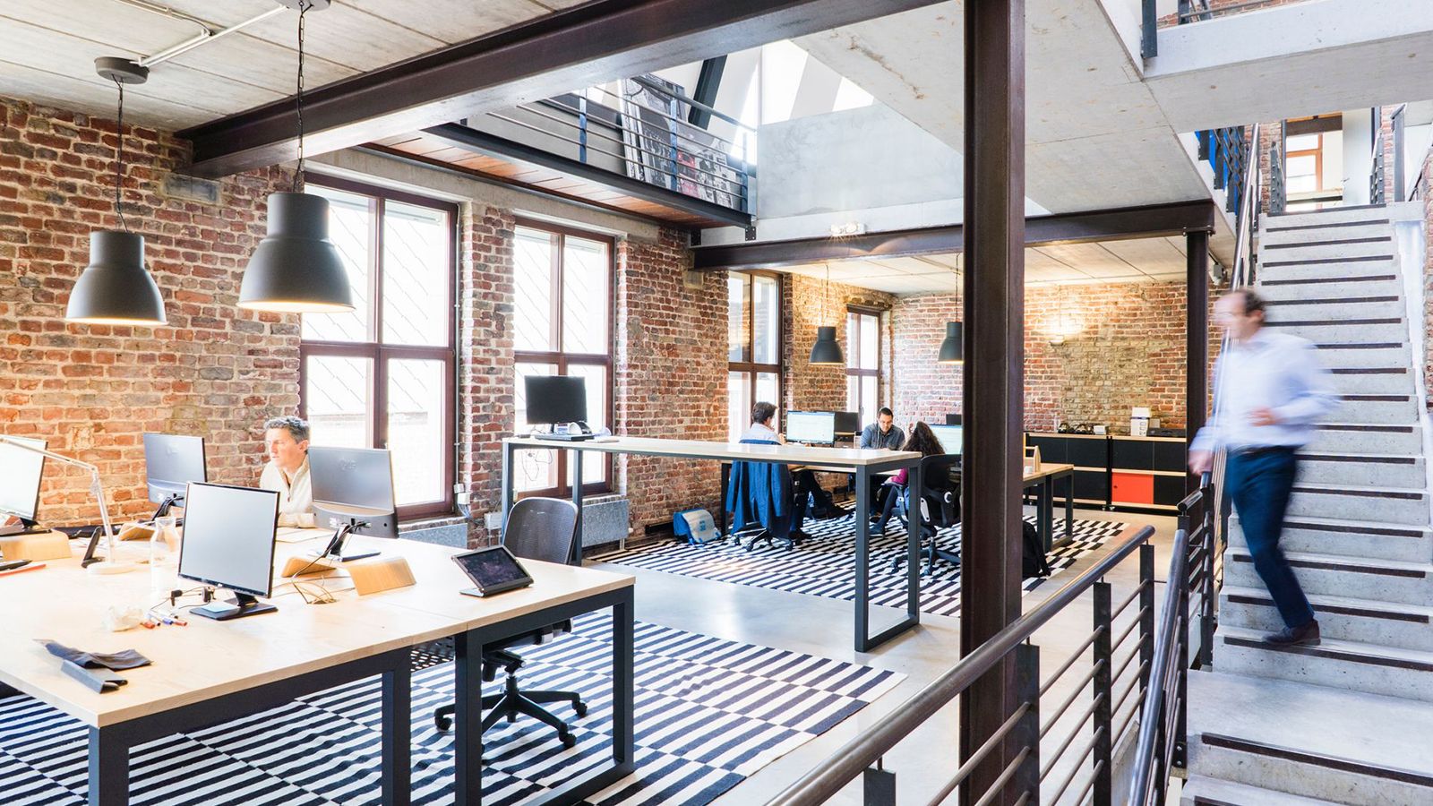 An open-plan office in a converted brick loft, with people working at desks and a staircase leading to a mezzanine.