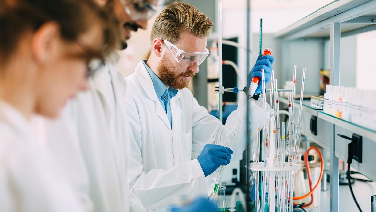Scientists in lab coats and safety glasses working with pipettes and test tubes in a research laboratory.