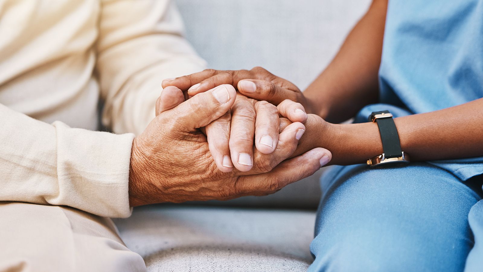 A caregiver in scrubs gently holds the hands of an older adult in a comforting gesture.