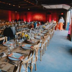 long farm tables set for dinner, orange napkins, sunflower centerpieces, and red uplighting, "Table Thirteen" sign in foreground.