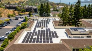 Aerial view of a commercial building with solar panels on its roof, with trees and water in the background